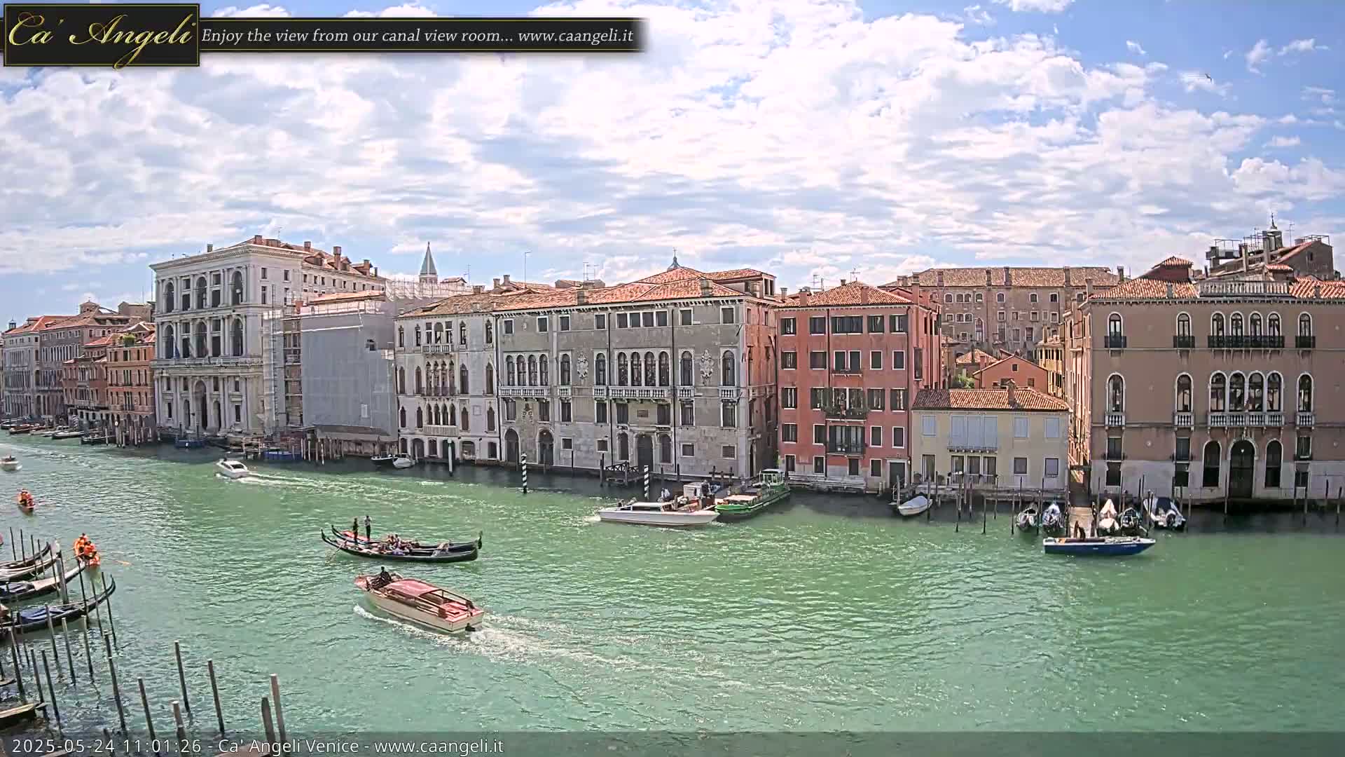 A sunny day in Venice shows a wide canal with gondolas and motorboats passing by colorful buildings lining the waterway.