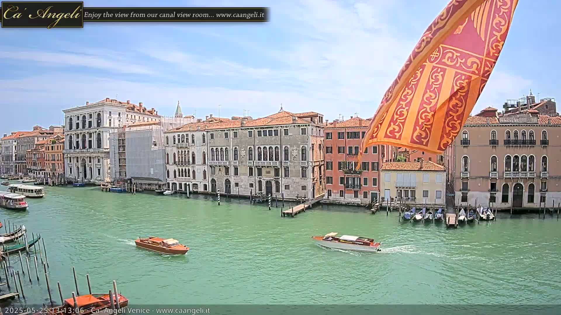 A canal in Venice, under a mostly sunny sky, is lined with historic buildings and several boats, with one orange and gold flag in the upper right corner.