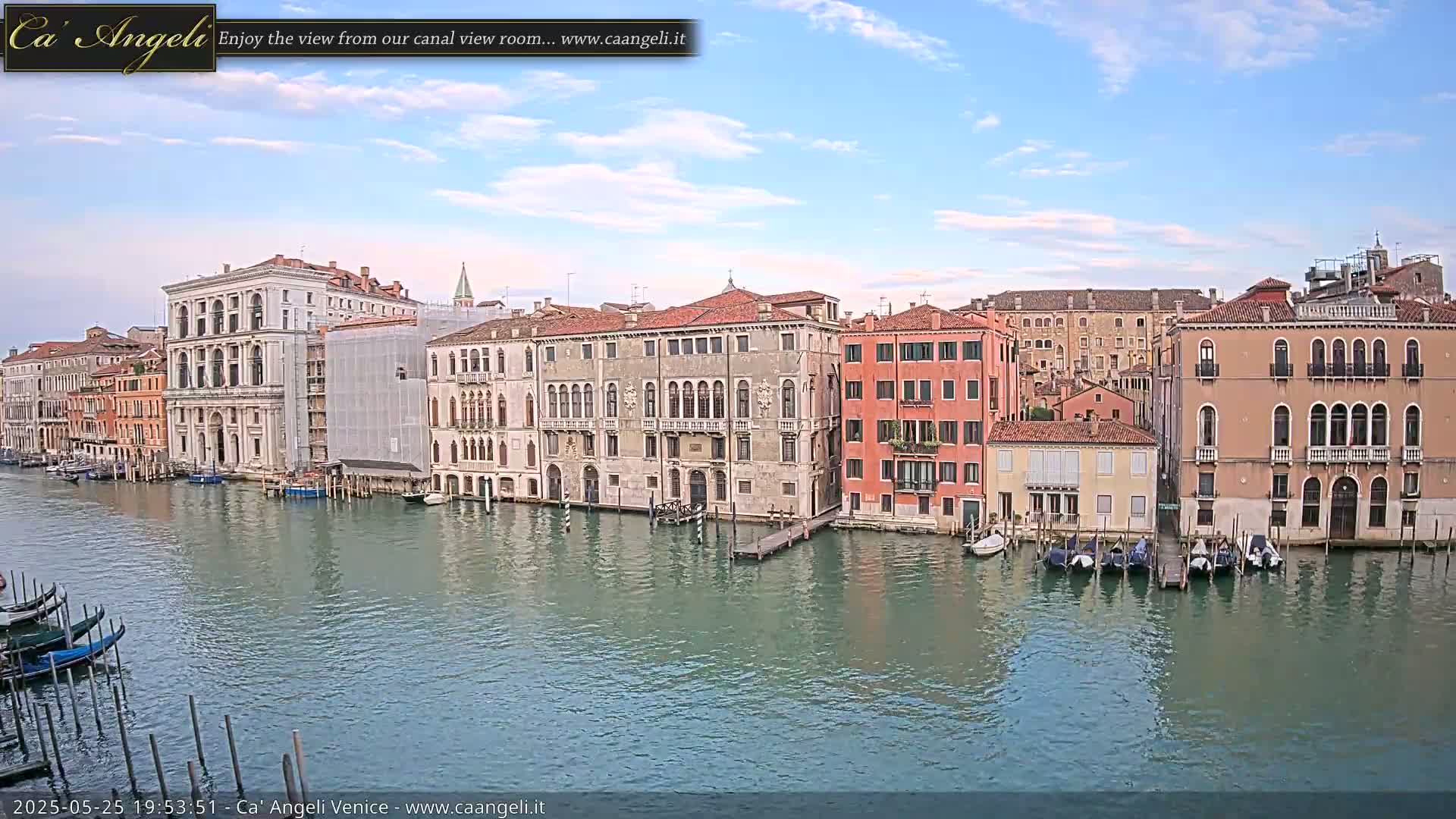 A canal in Venice, under a mostly clear sky with a few clouds, is lined with historic buildings and gondolas moored along the banks.