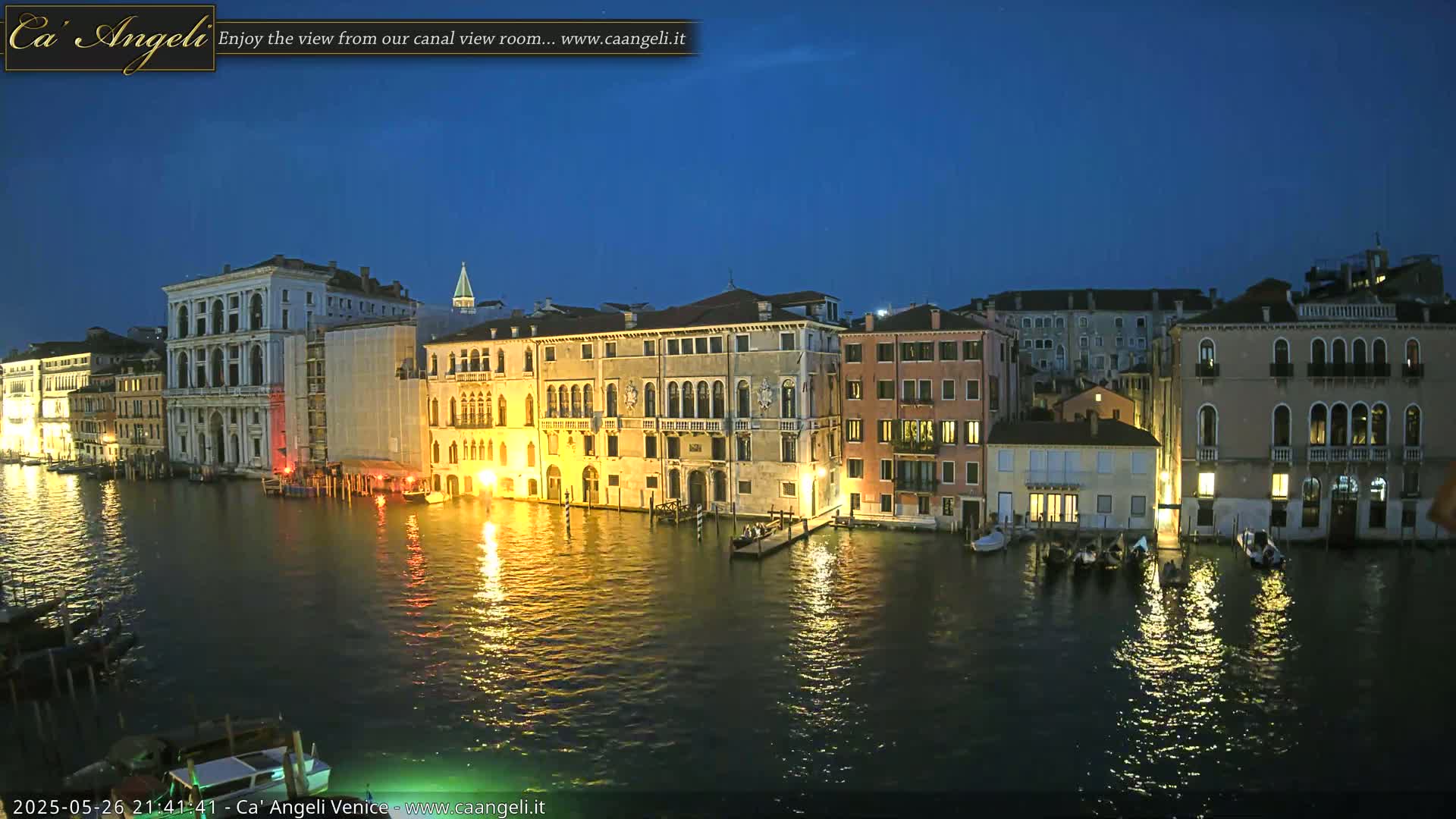 A nighttime view of a canal in Venice, Italy, shows several illuminated buildings lining the waterway, under a clear, dark blue sky.