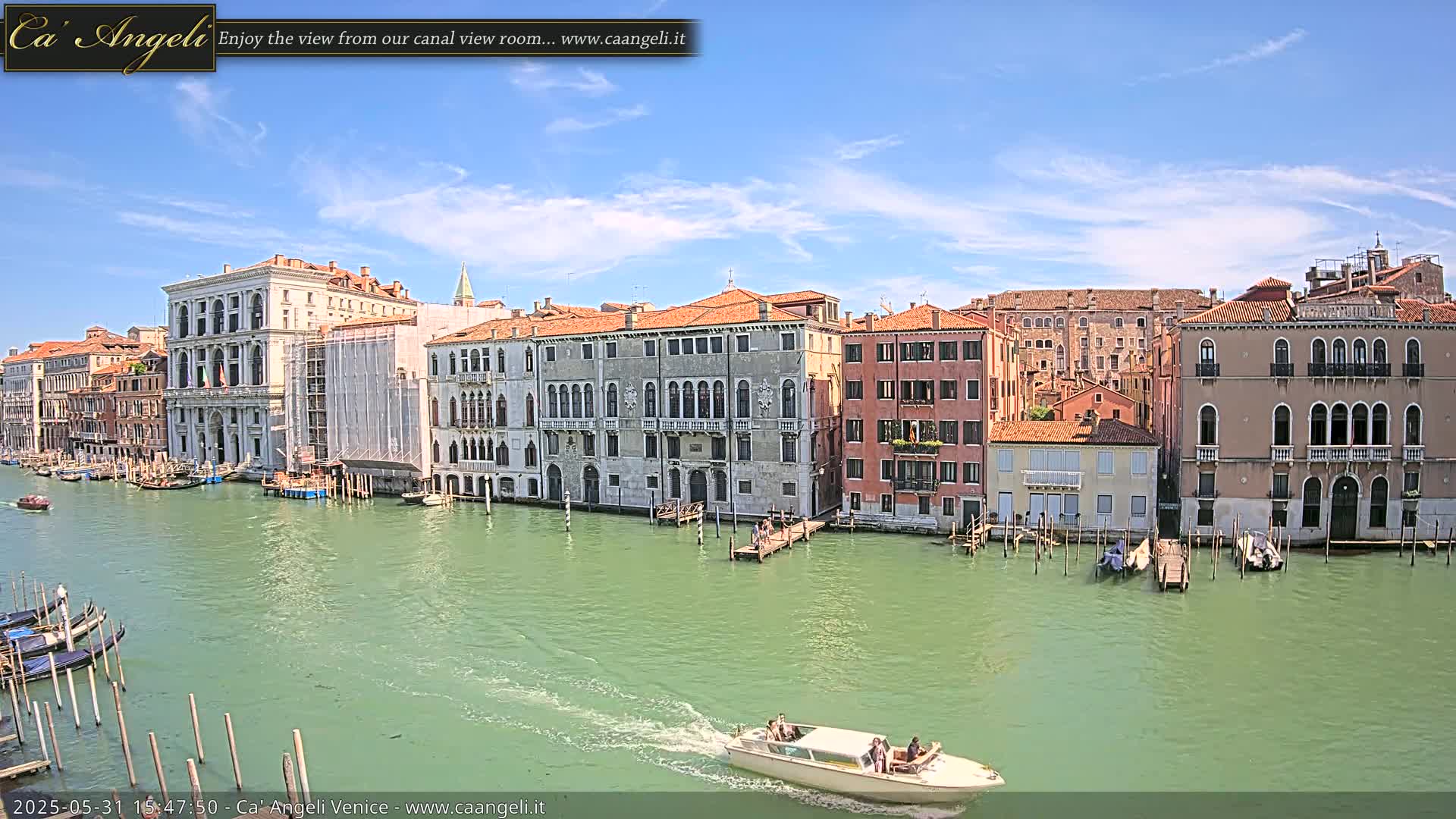 A sunny day in Venice shows a canal with several moored gondolas and a motorboat passing by; numerous buildings line the waterway.