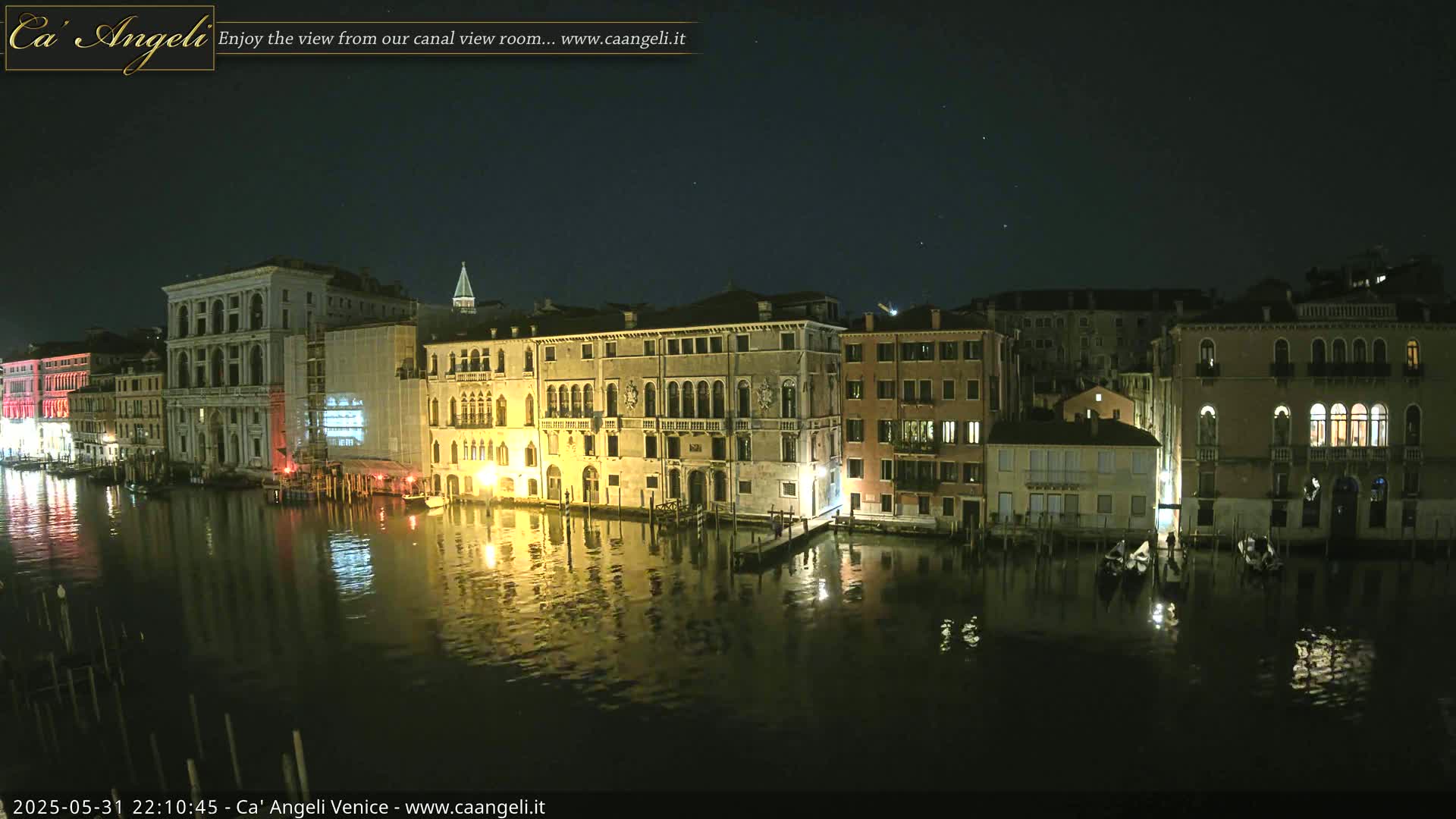 A nighttime view of a canal in Venice, with illuminated buildings lining the waterway and their lights reflecting in the calm water under a clear, starry sky.