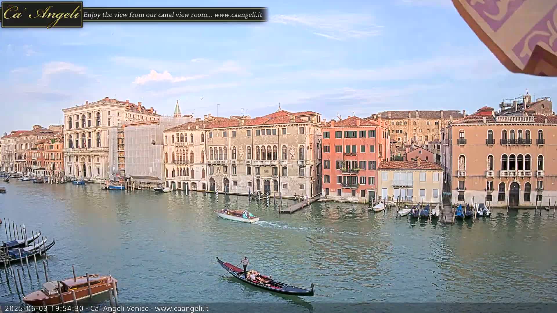 A gondola glides along a canal in Venice, Italy, under a mostly clear sky, past numerous buildings and moored boats.