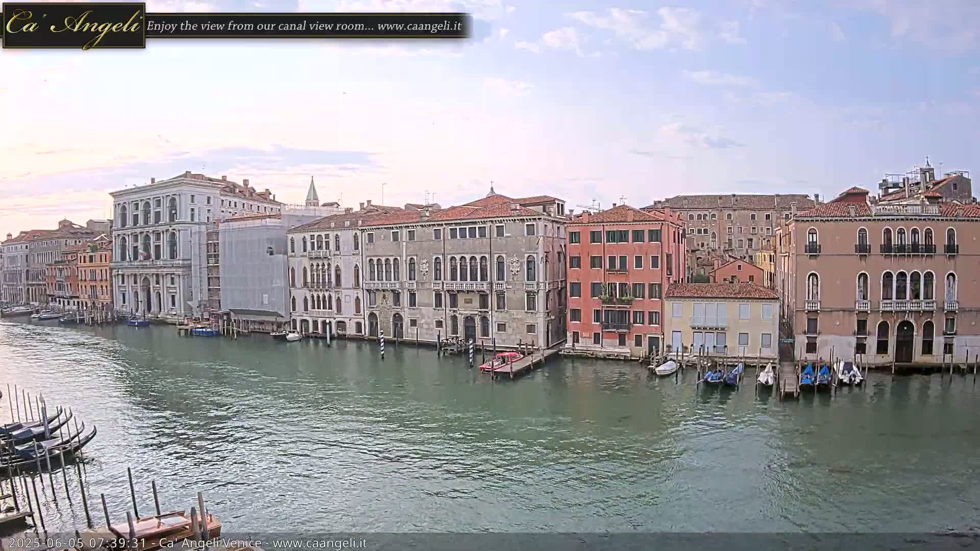 A canal in Venice, under a partly cloudy sky, is lined with several multi-story buildings, some of which are under construction, with boats moored along the waterway.