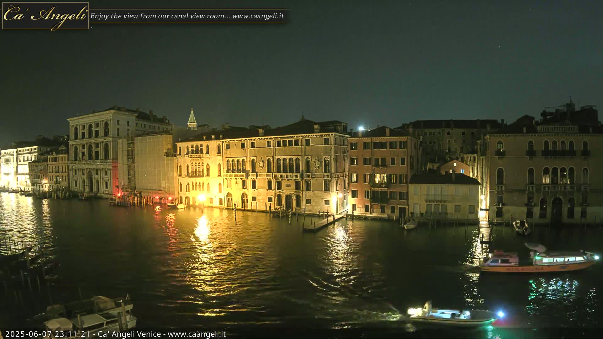 A nighttime view of illuminated buildings lining a canal in Venice, with several boats on the water under a clear, dark sky.