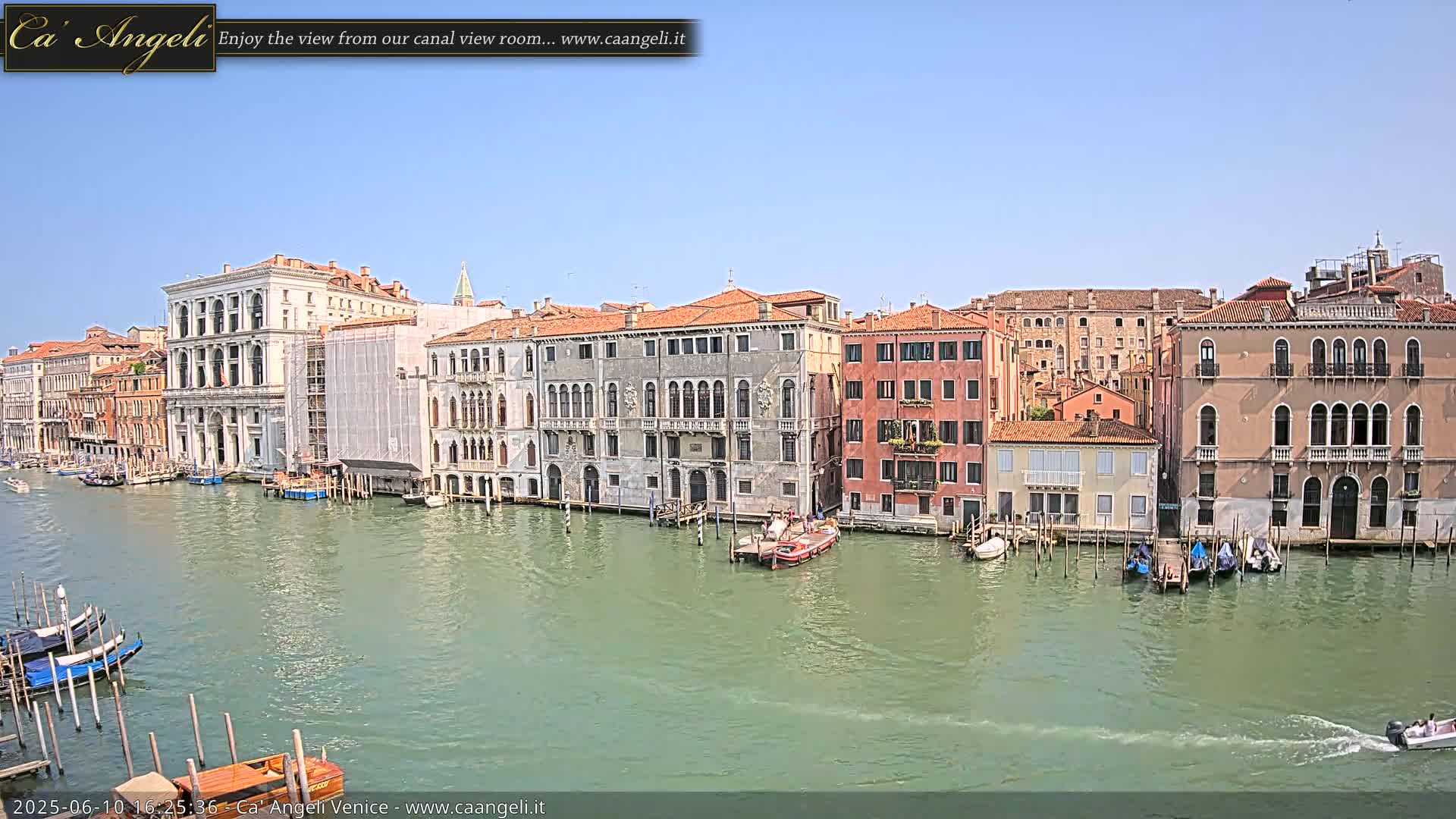 A sunny day in Venice shows a canal lined with historic buildings, boats moored along the banks, and a motorboat speeding across the water.