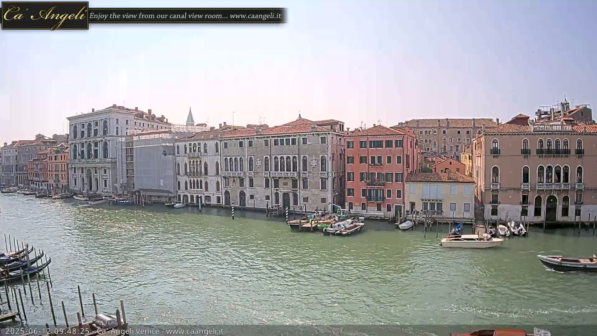 A sunny day in Venice shows several canal-side buildings, boats moored along the waterway, and gondolas at a dock.