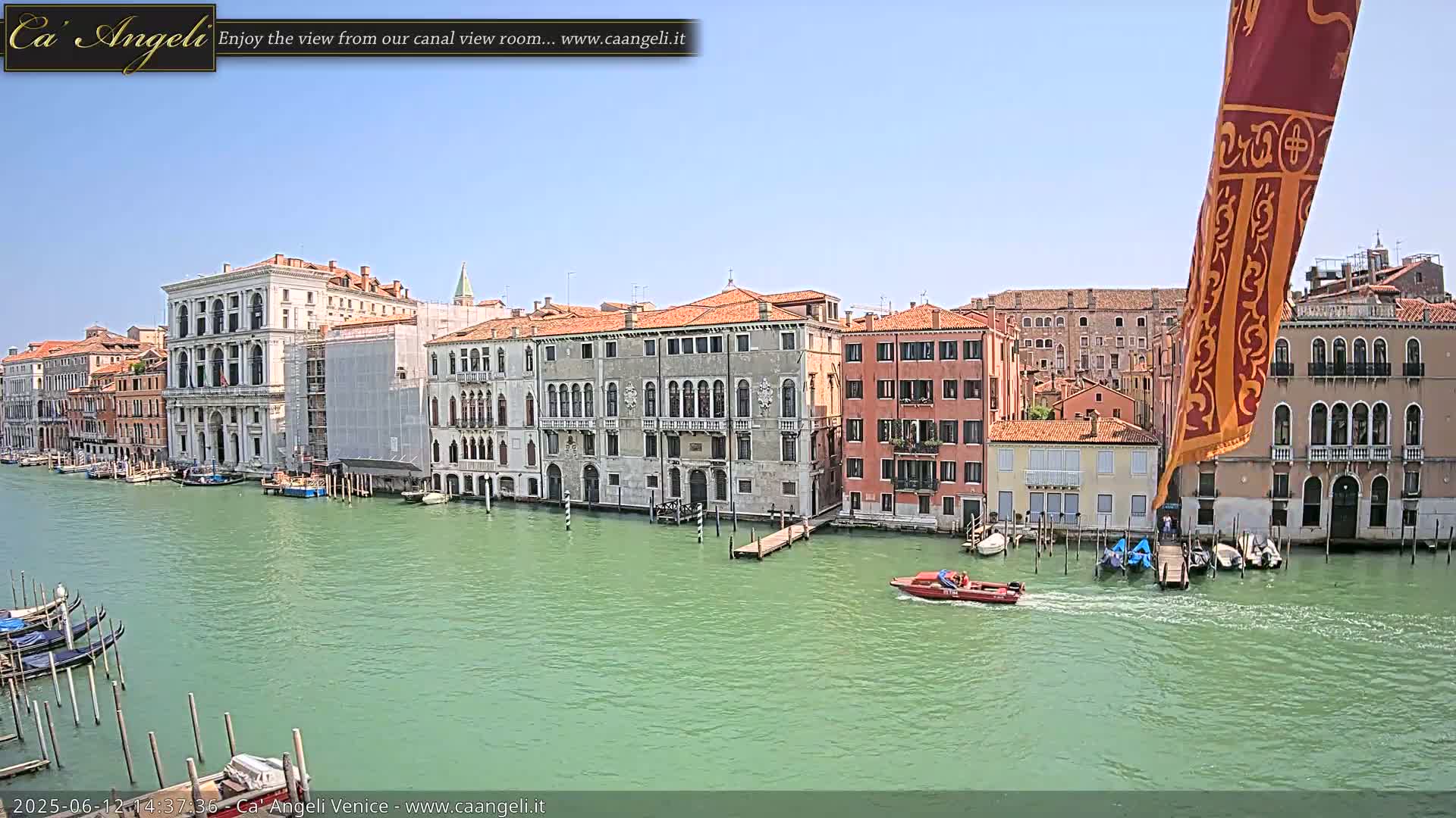 A sunny day in Venice shows a canal with several gondolas and a motorboat passing by numerous buildings along the waterway.