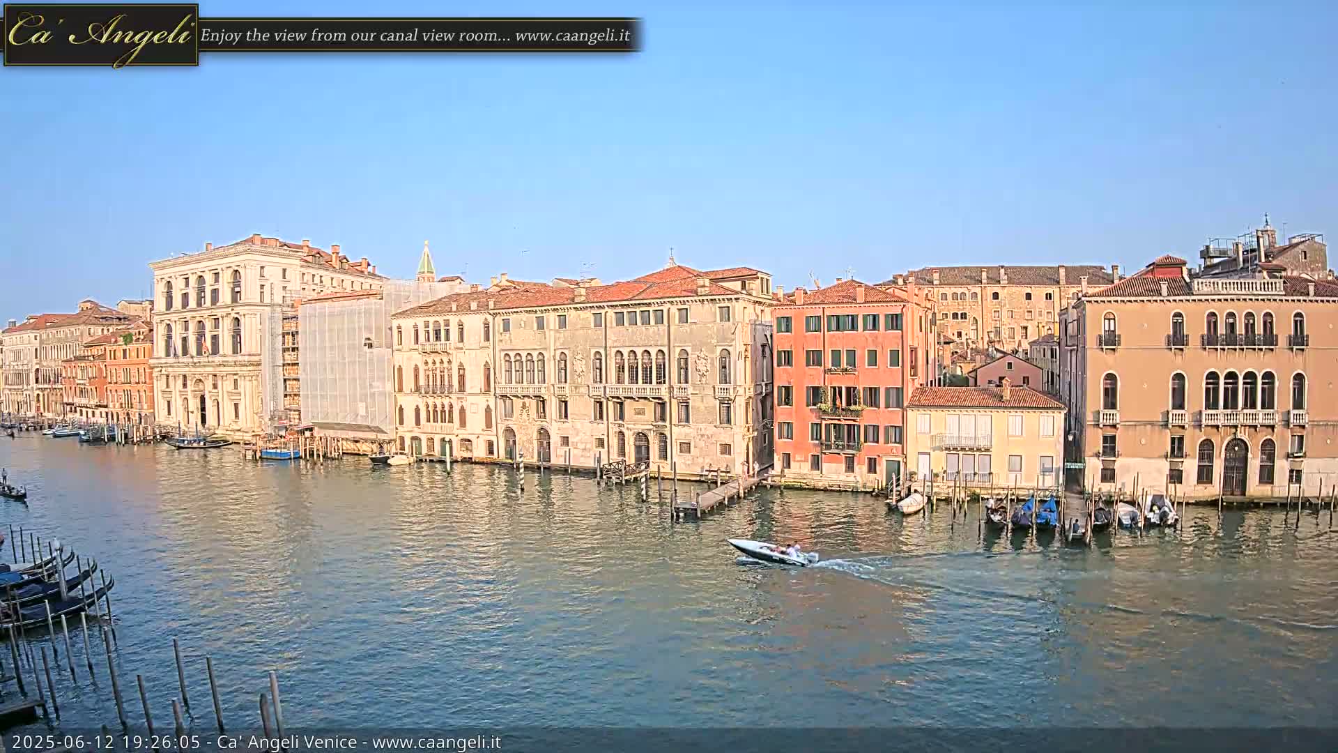 A sunny day in Venice shows a canal with several buildings lining its edges, gondolas moored along the shore, and a motorboat speeding across the water.