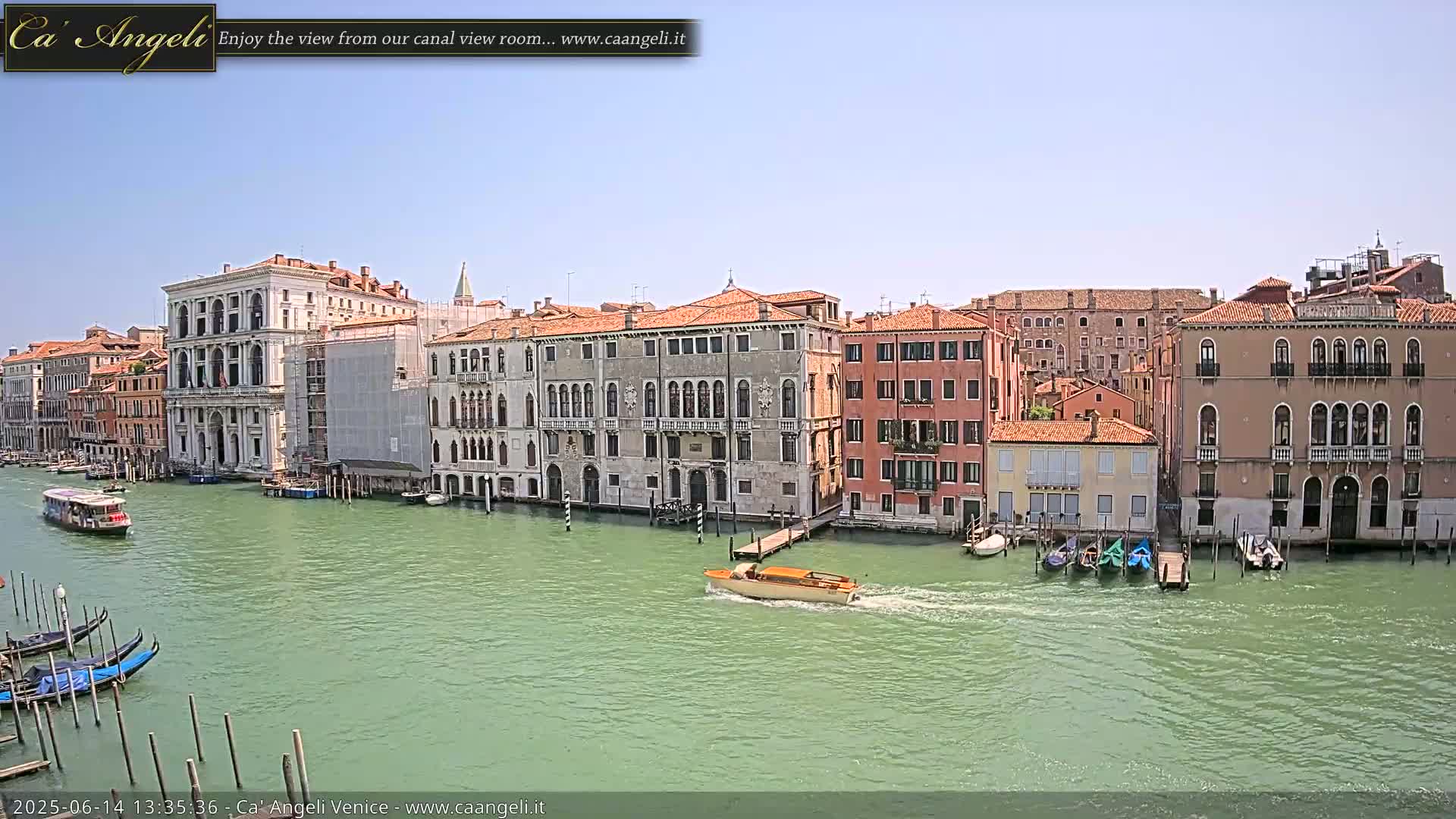 A sunny day in Venice shows a canal with several gondolas, a motorboat, and numerous buildings lining the waterway.