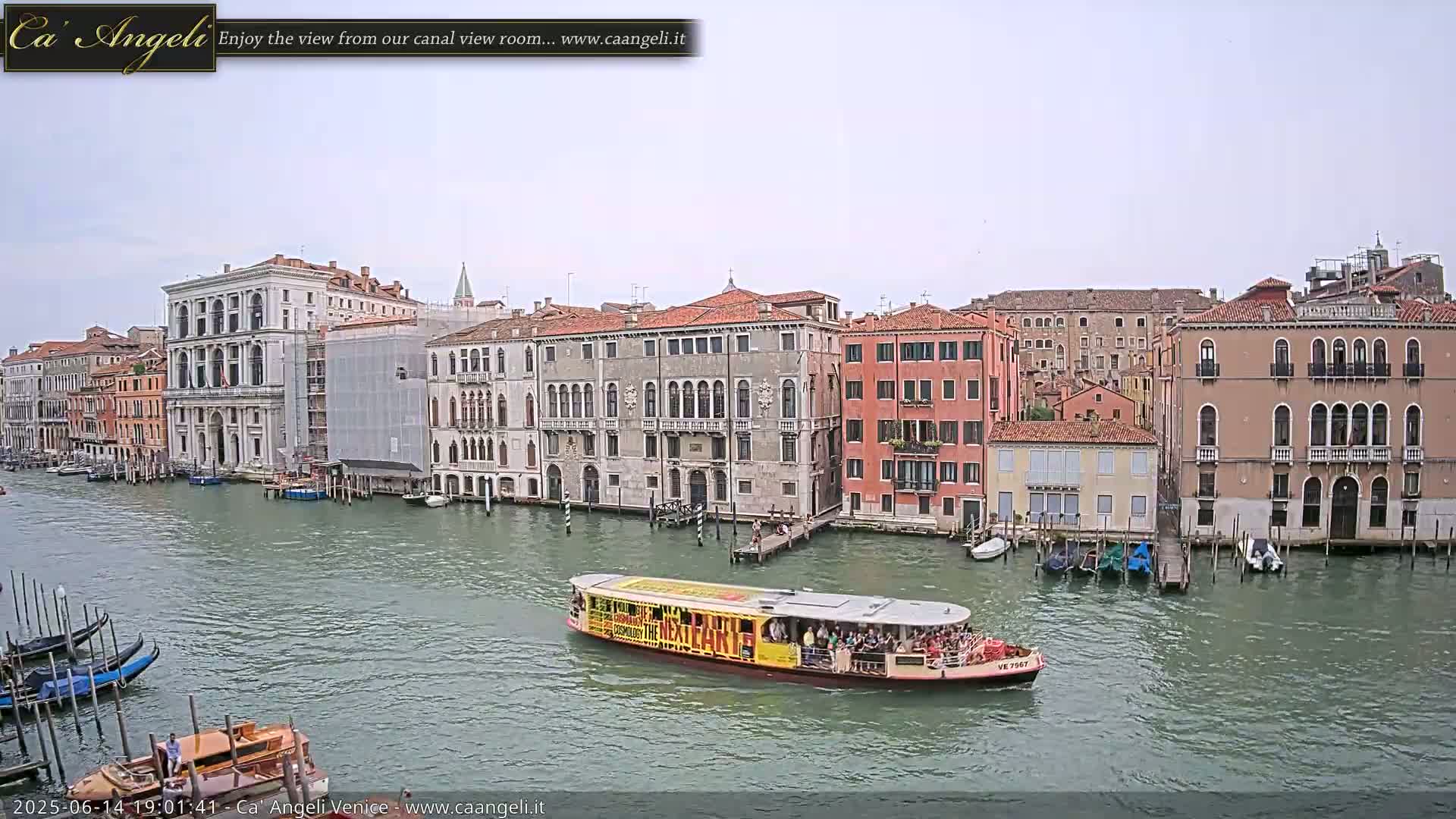 A canal in Venice, under an overcast sky, shows several gondolas moored along the banks, alongside a passenger boat traveling on the water, and numerous buildings lining the waterway.