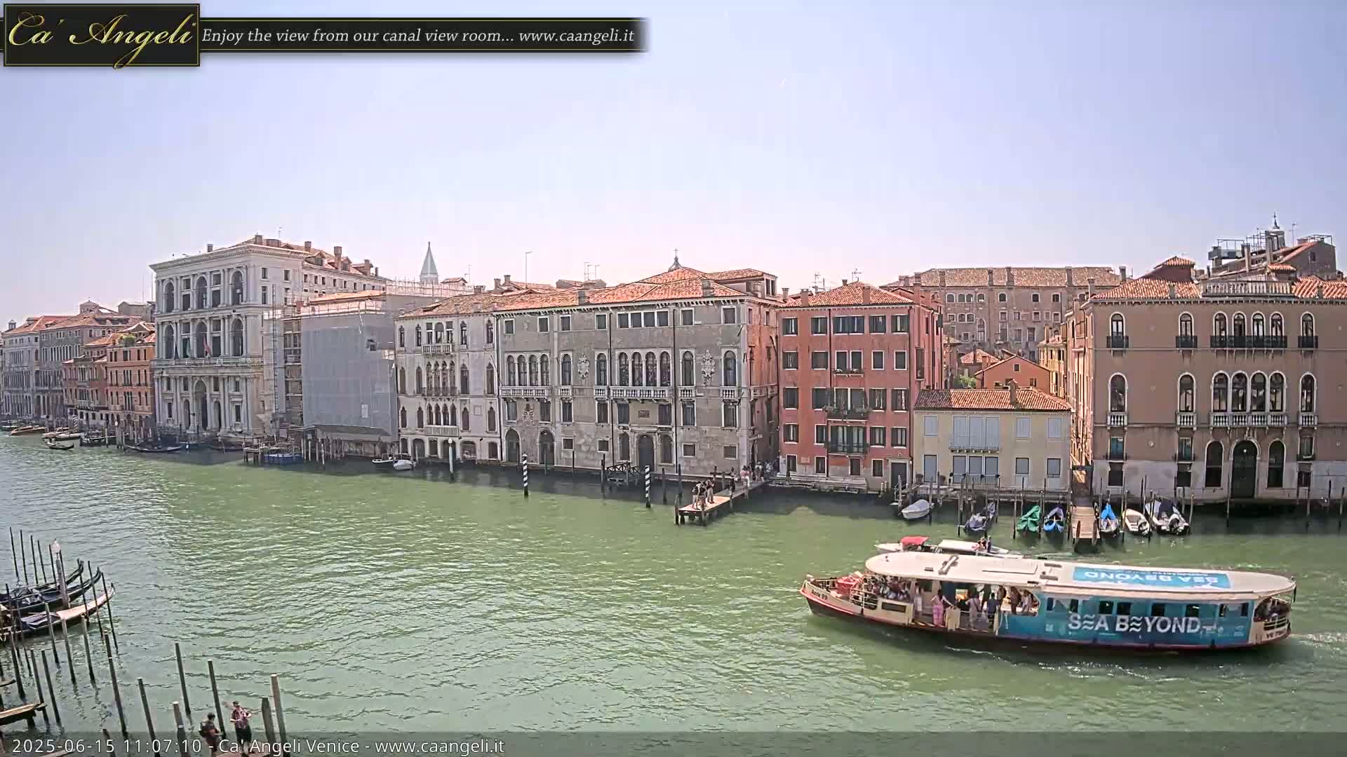 A sunny day in Venice shows a canal with several buildings lining its banks, a water taxi with passengers, and gondolas moored at the side.