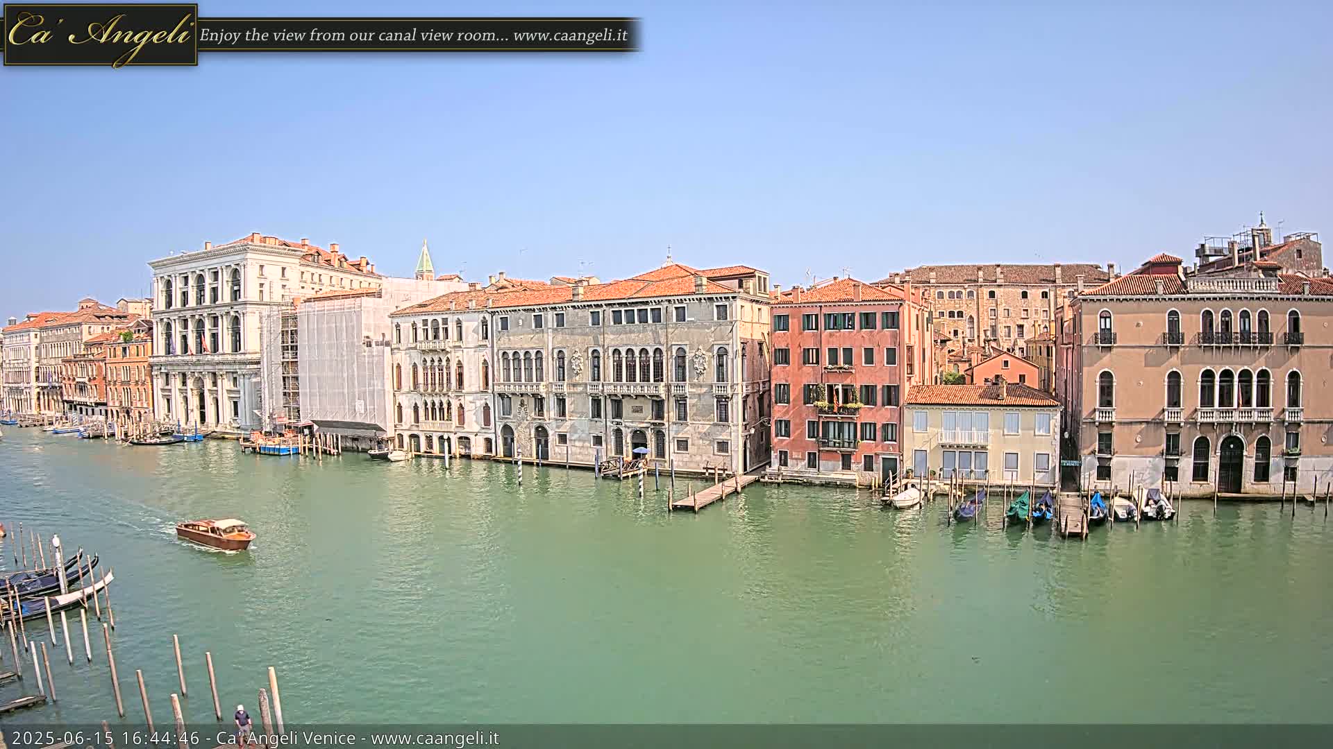 A sunny day in Venice shows a wide canal with several gondolas and small motorboats moored along the waterfront,  lined with numerous historic buildings.
