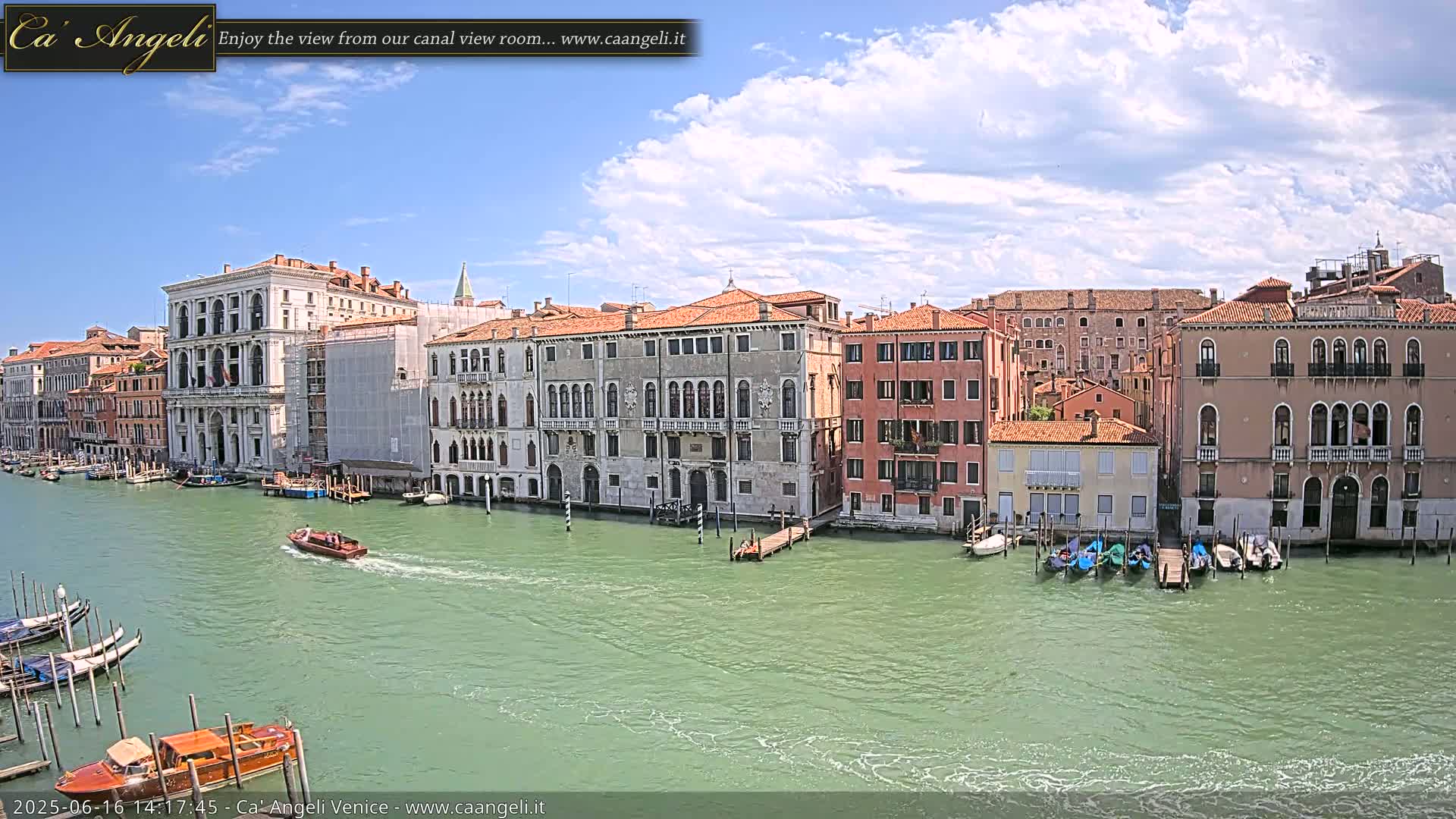 A sunny day in Venice shows a canal with several gondolas and boats moored alongside a row of buildings.