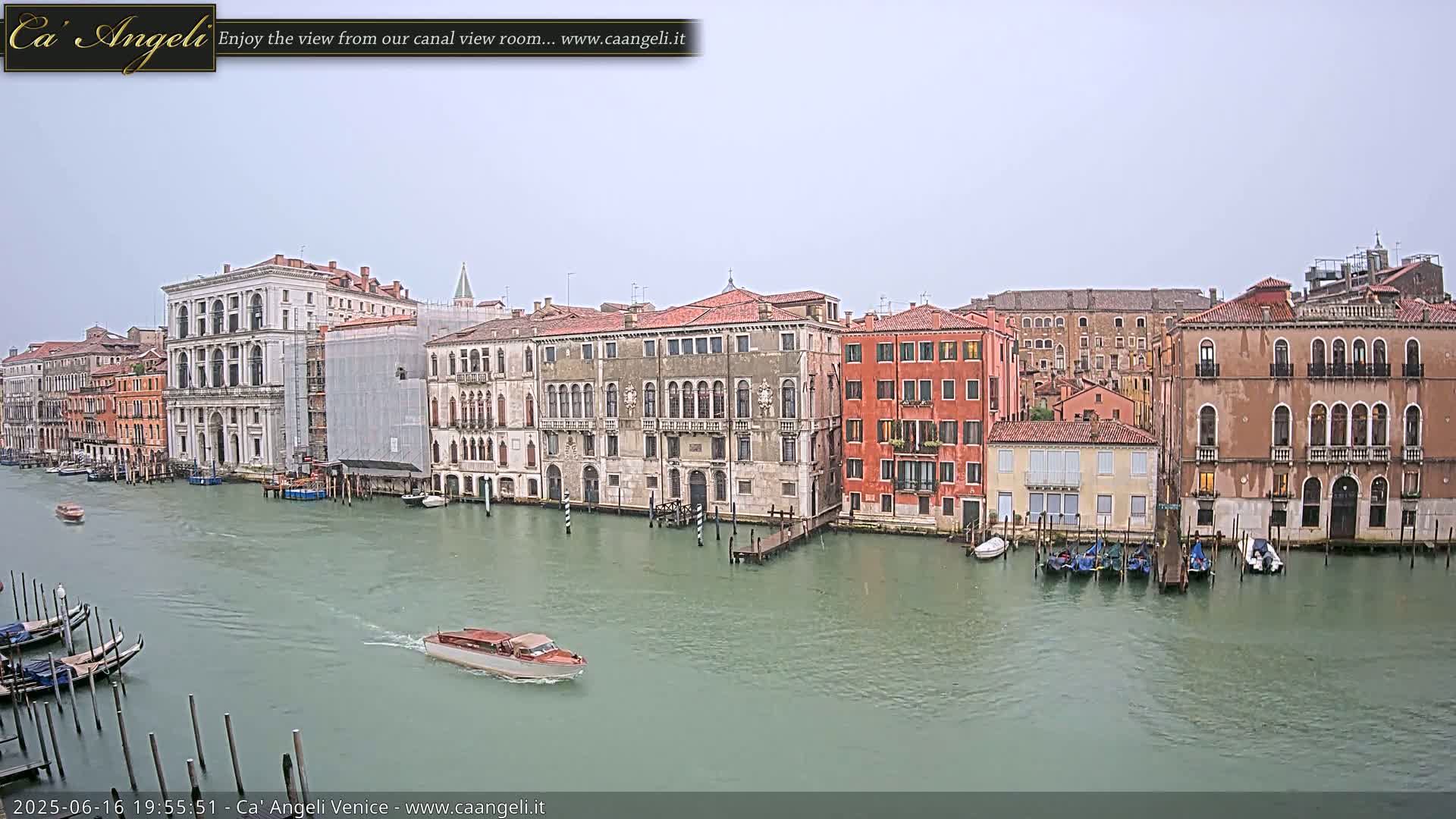 A canal in Venice, Italy, on an overcast day shows several historic buildings lining the waterway, with gondolas and a motorboat on the water.