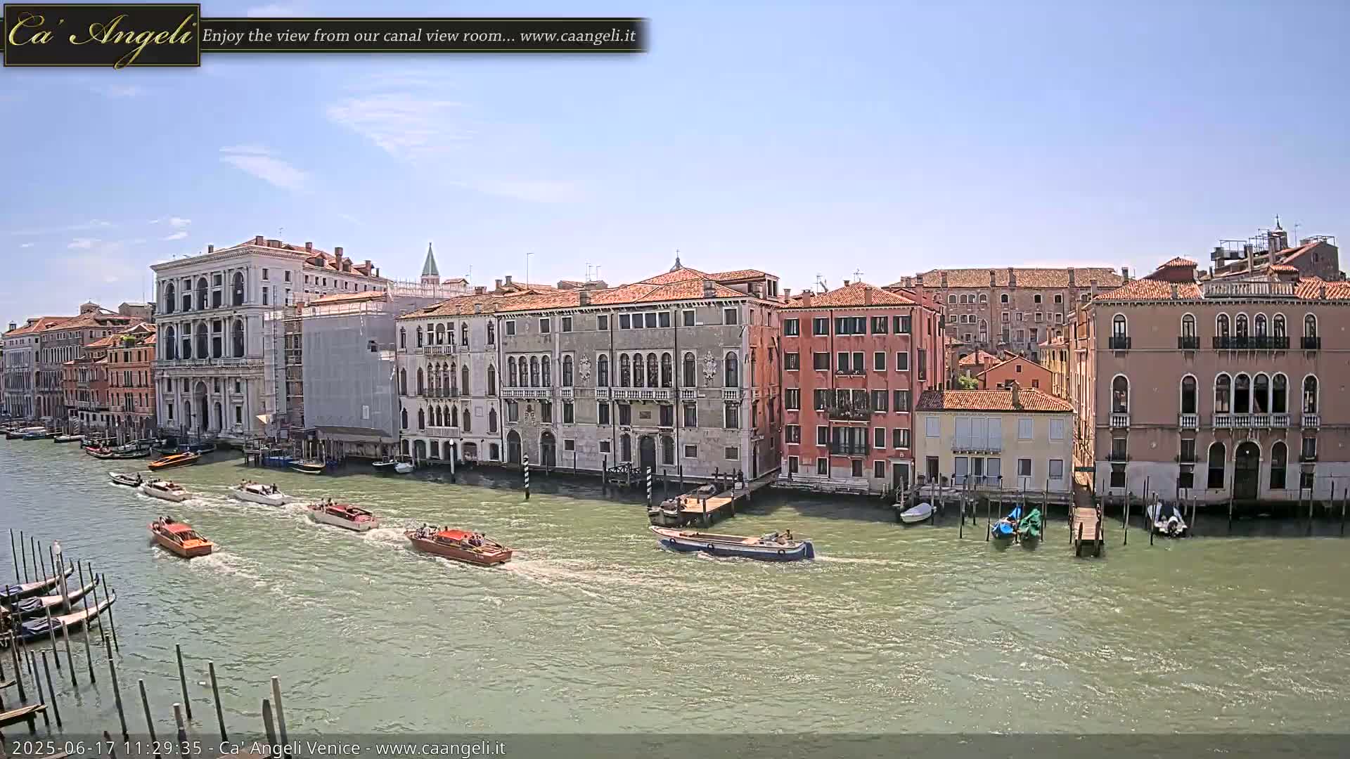 Under a clear blue sky, several motorboats travel down a canal lined with historic Venetian buildings.