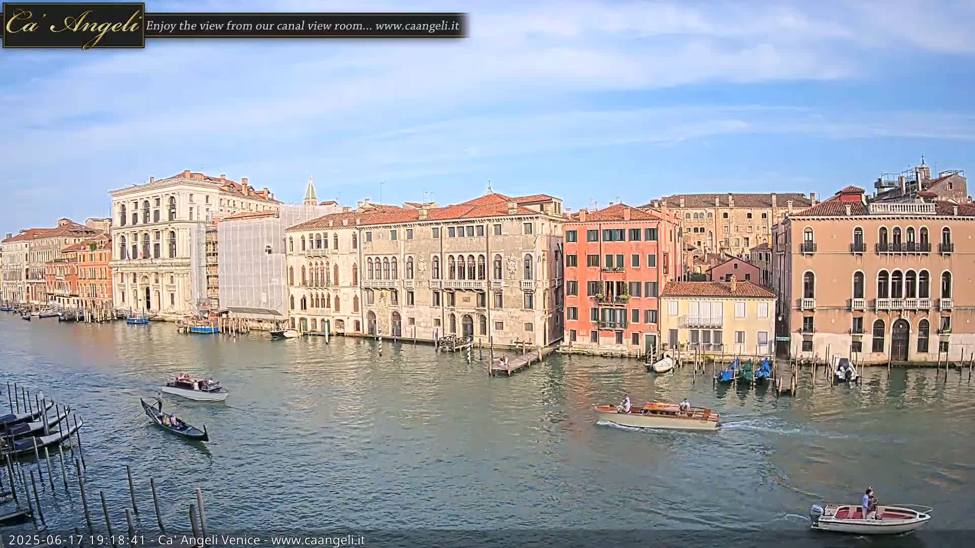 A sunny day in Venice shows several gondolas and motorboats on a canal alongside tall, historic buildings.