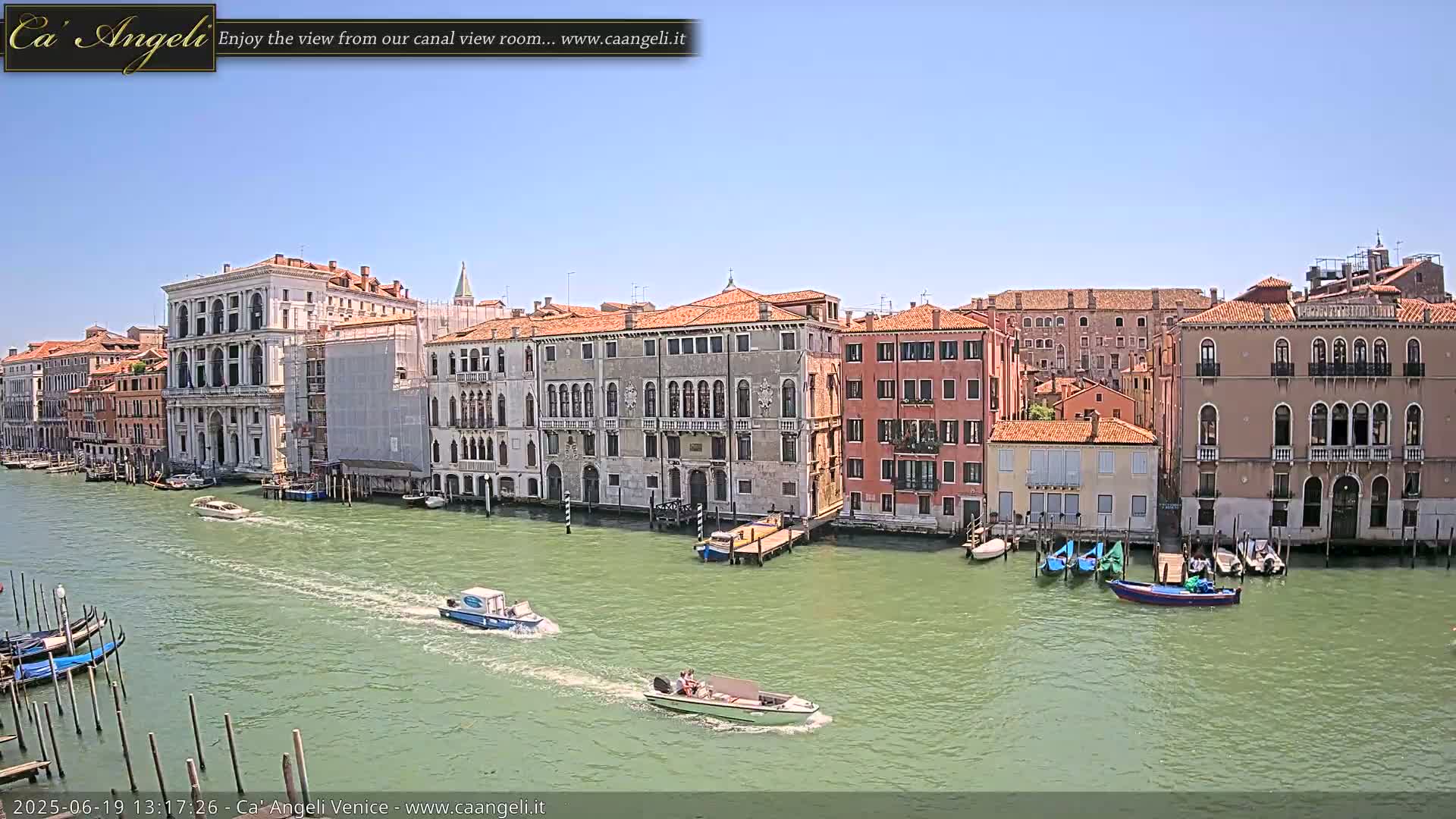 A sunny day in Venice shows several canal-side buildings, gondolas moored at the water's edge, and several motorboats traveling on the green-tinged canal.