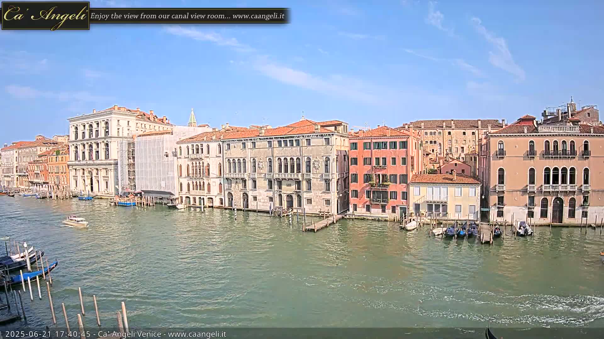 A sunny day in Venice shows a canal lined with historic buildings, gondolas moored alongside, and a few boats moving on the water.
