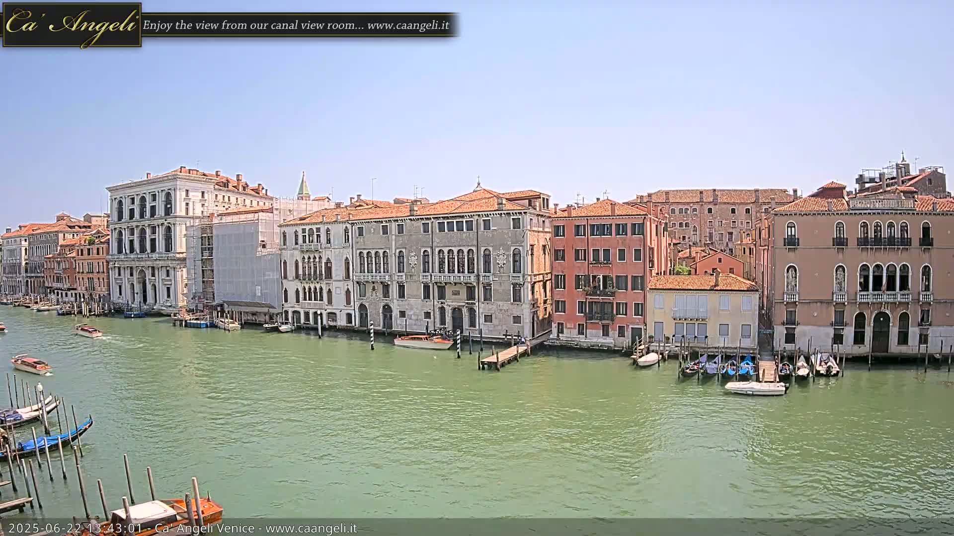 A canal in Venice, under a clear blue sky, is lined with historic buildings and several boats are moored along the waterway.