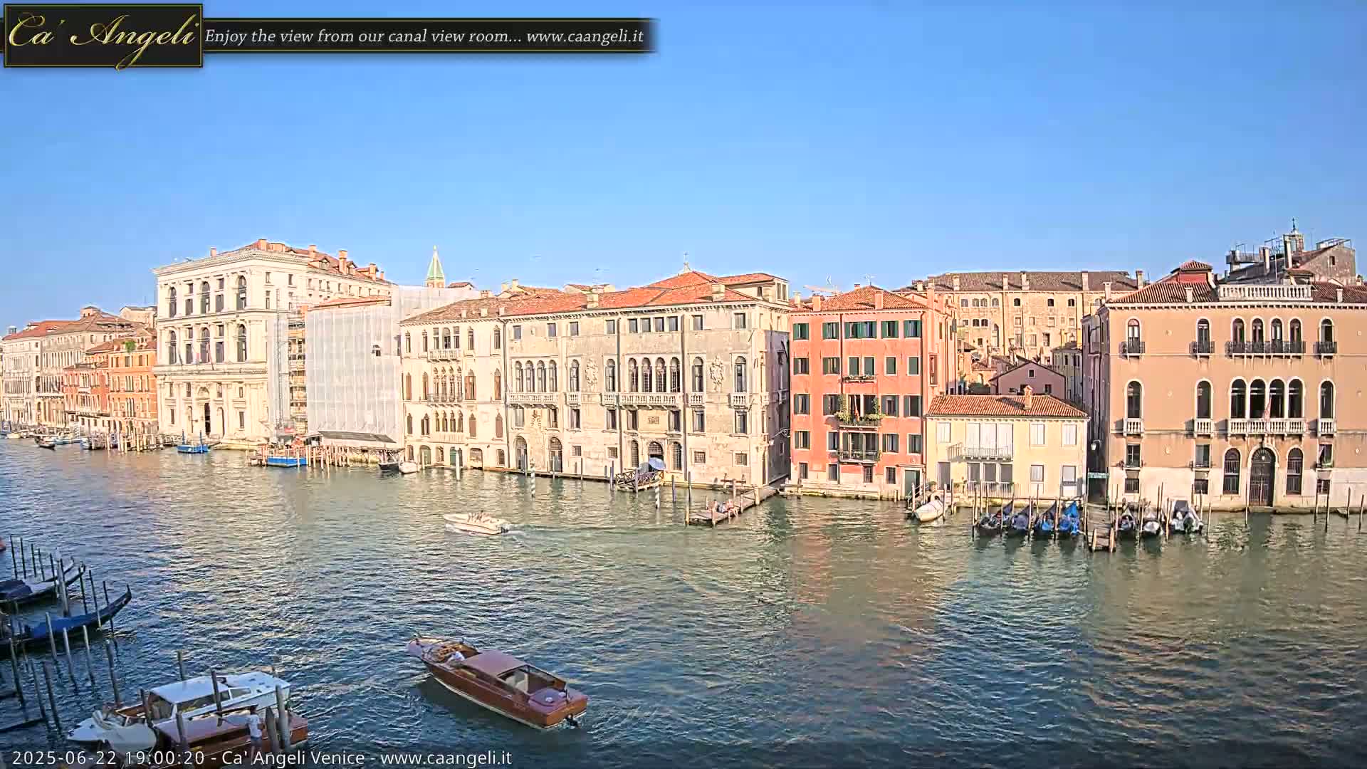 A sunny day in Venice shows several canal-side buildings, gondolas moored at a dock, and a motorboat on the water.
