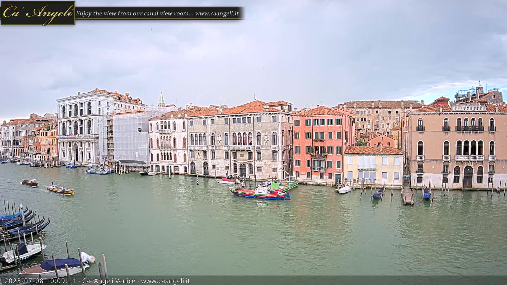 A canal in Venice, under an overcast sky, is lined with several multi-story buildings, with boats moored along the edges and a few others traveling the waterway.