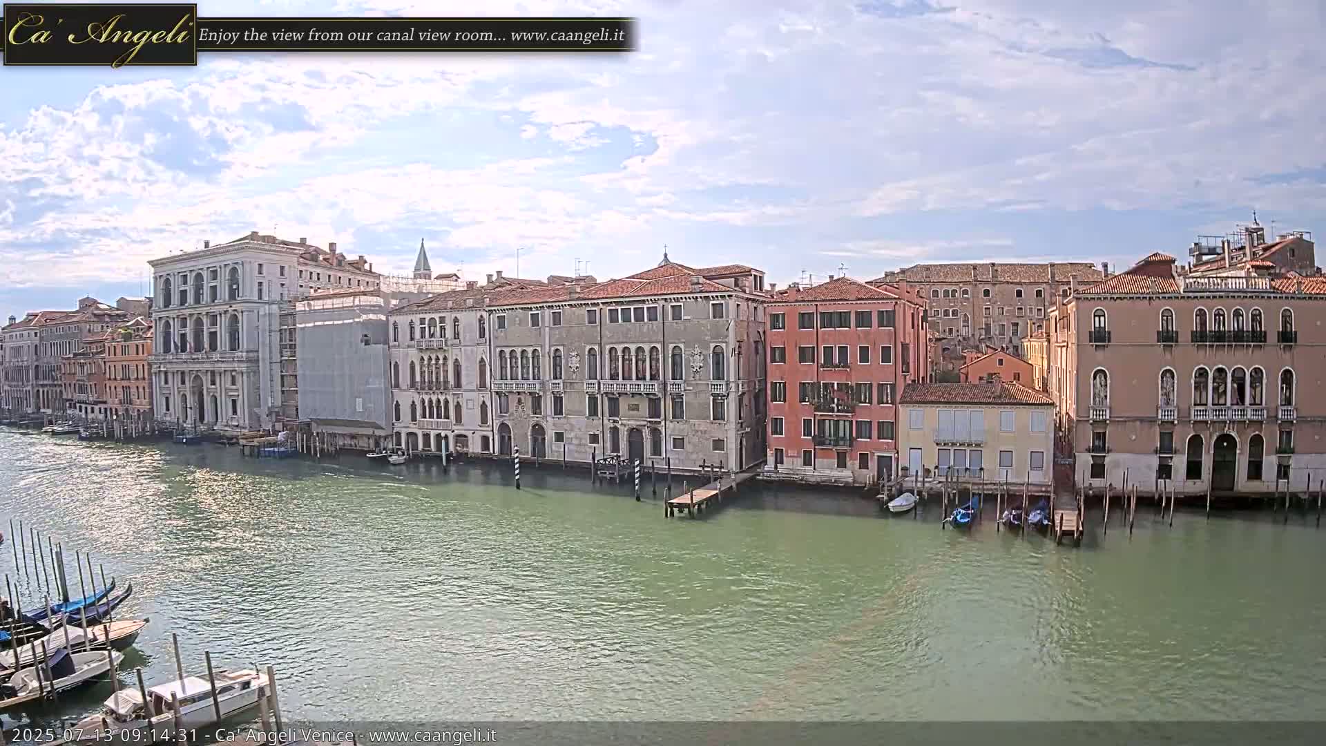 A canal in Venice, under a partly cloudy sky, is lined with numerous historic buildings, with gondolas and small boats moored along the waterfront.
