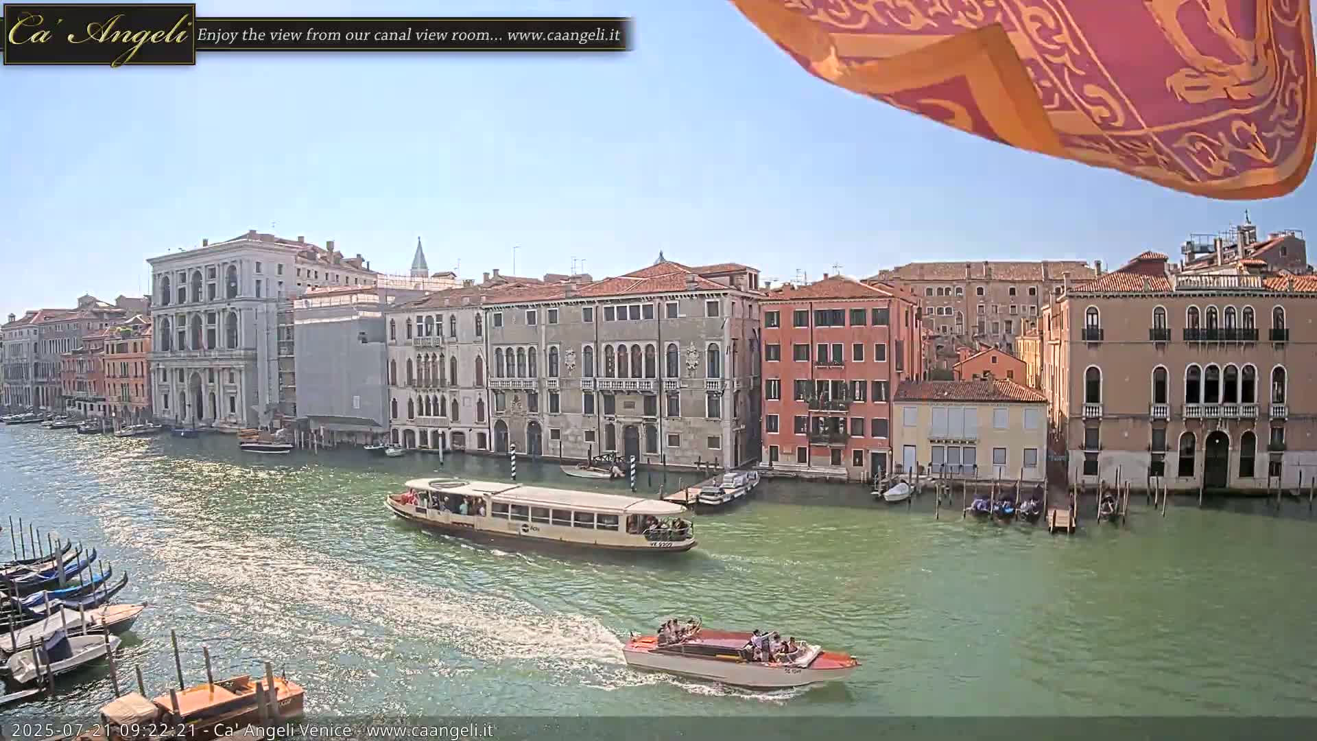 A sunny day in Venice shows a canal with several boats, including a water bus and gondolas, passing by buildings along the waterfront.