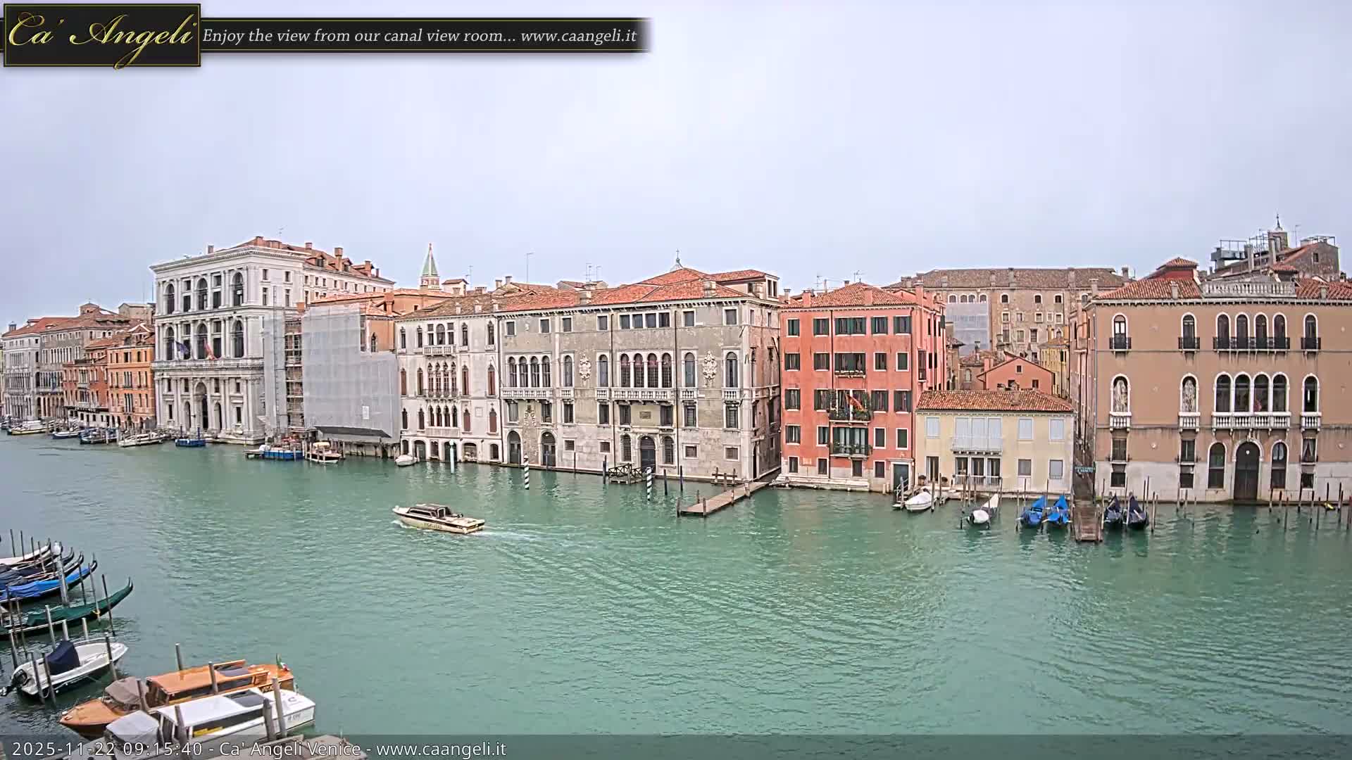 A wide, elevated view reveals a Venetian canal, its banks densely packed with colorful historic buildings and numerous moored boats, as a small motorboat cruises across the greenish-blue water under an overcast sky.