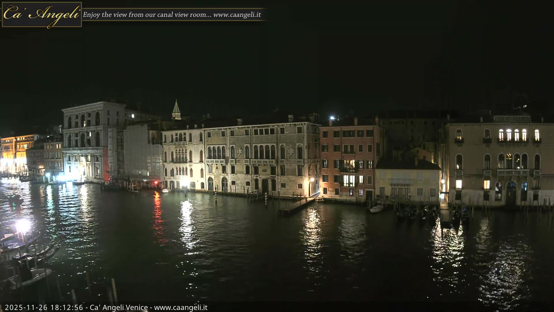 A wide, elevated view reveals a Venetian canal, its banks densely packed with colorful historic buildings and numerous moored boats, as a small motorboat cruises across the greenish-blue water under an overcast sky.