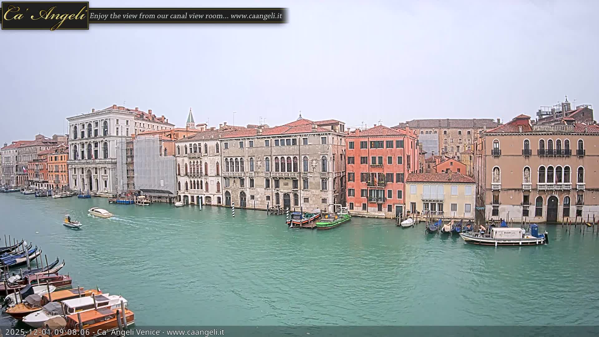 A wide view of the Grand Canal in Venice on an overcast day, showcasing historic buildings of varying colors and architectural styles lining the turquoise water, with numerous boats and gondolas docked and some motorboats actively traversing the canal.