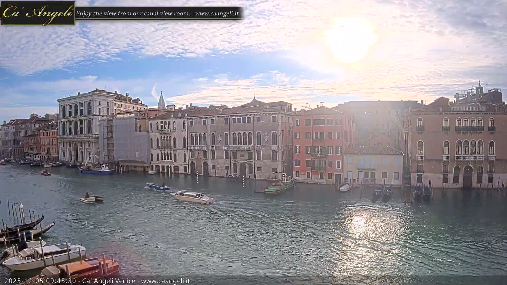 A wide canal, bustling with various boats, winds between numerous multi-story buildings with varied architecture under a bright, partly cloudy sky.