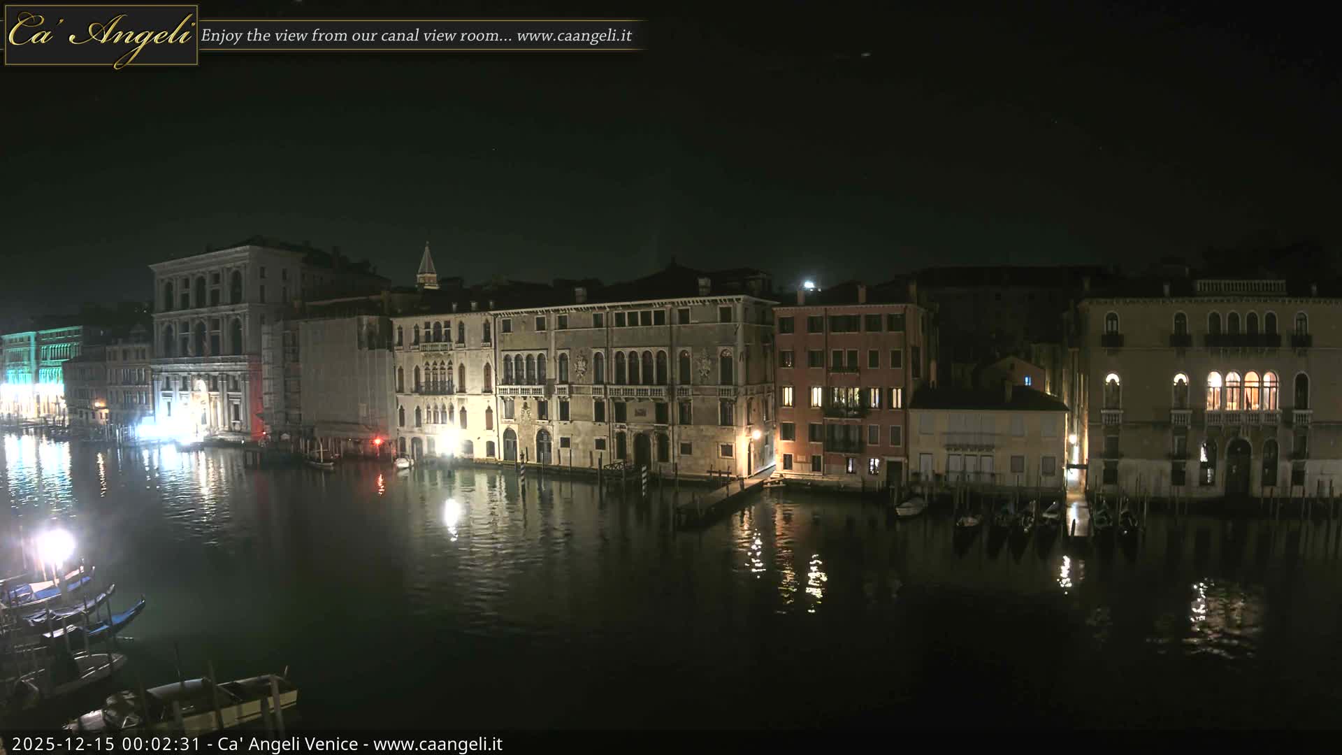 A wide canal, bustling with various boats, winds between numerous multi-story buildings with varied architecture under a bright, partly cloudy sky.