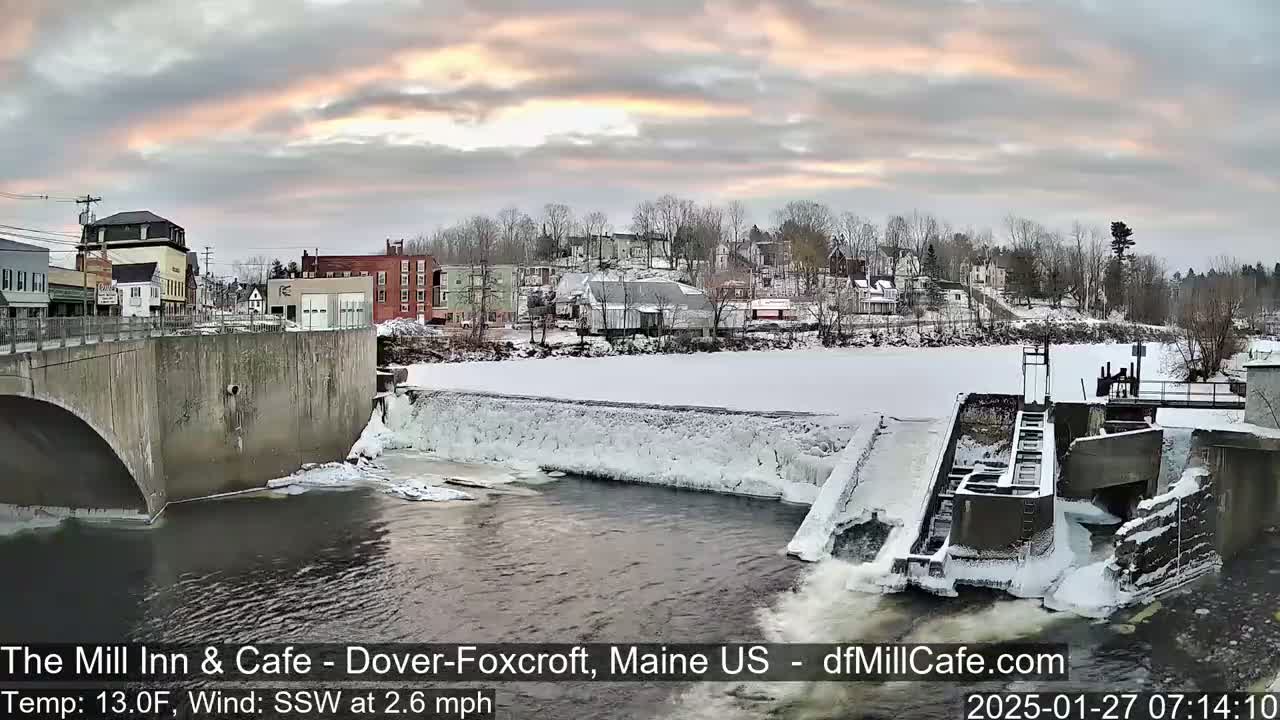 A partially frozen river flows under a bridge past a dam in a snow-covered town during a sunrise with a temperature of 13 degrees Fahrenheit and a south-southwest wind.