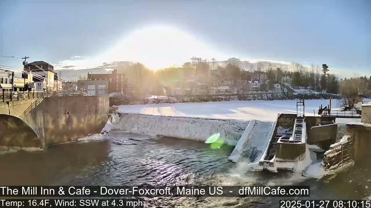 A partly sunny winter scene shows a river flowing past a snow-covered dam and bridge, with a town visible in the background.