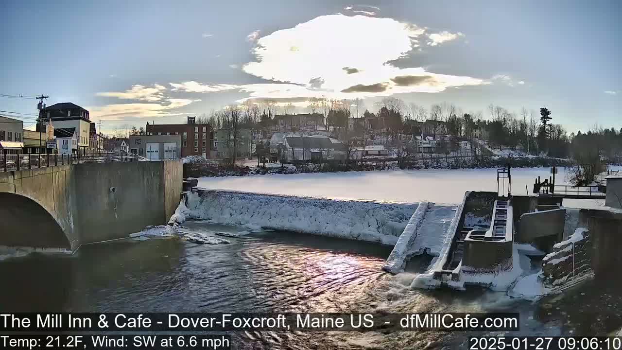 A partially frozen river flows past a dam and under a bridge in a snow-covered town, under a partly cloudy sky.