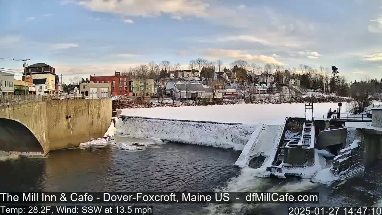 A partially frozen river flows past a dam and under a bridge in a snow-covered town, under a partly cloudy sky.