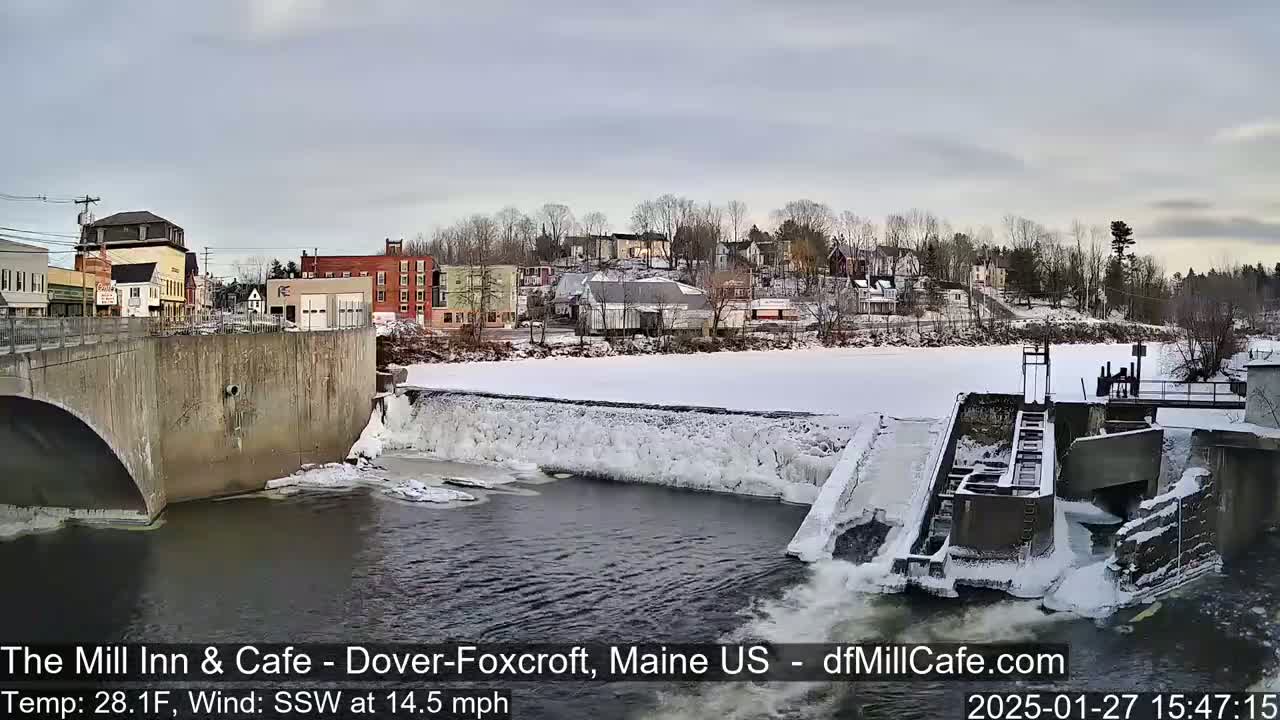 A partially frozen river flows past a dam and under a bridge in a small town, with snow covering the ground and buildings in the background under a cloudy sky.