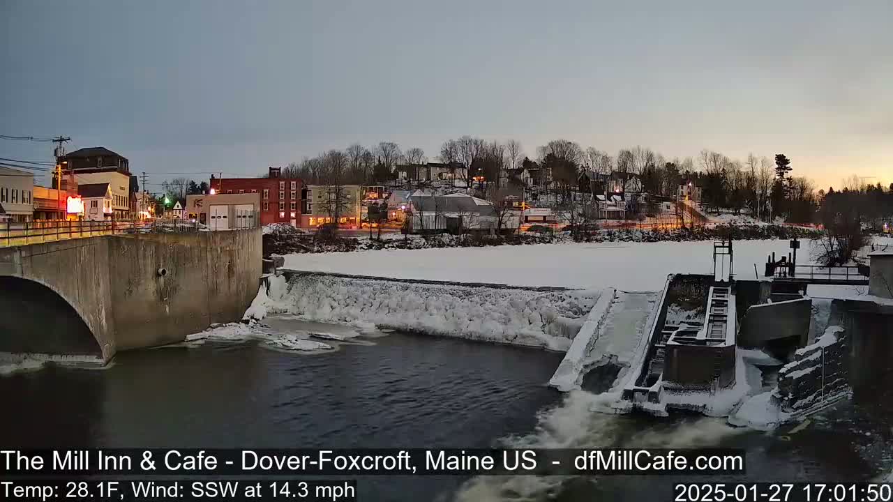 A snow-covered dam and partially frozen river flow past a town at twilight, with a cold wind blowing from the southwest.