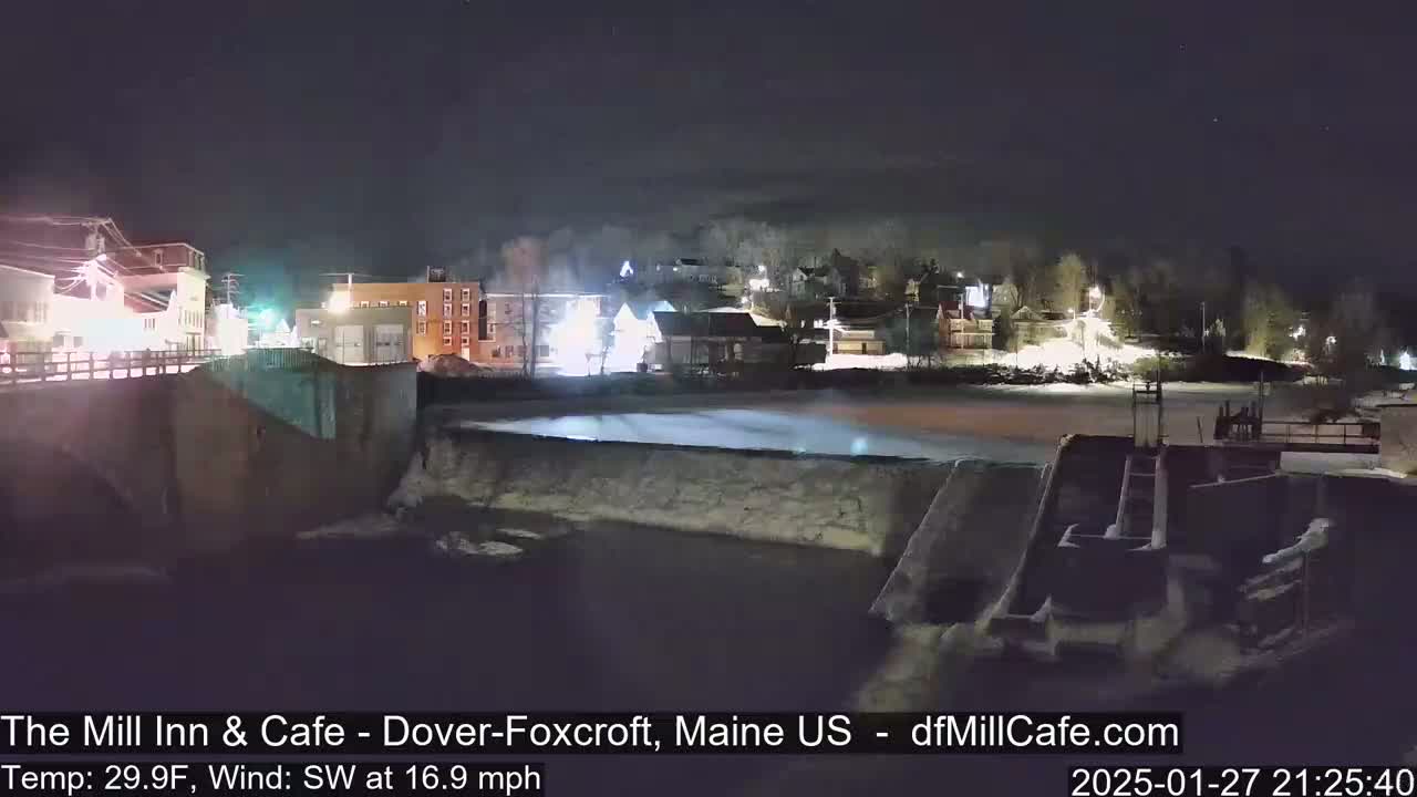 A nighttime view of a town, partially snow-covered, with a dam and river in the foreground under a clear, dark sky.