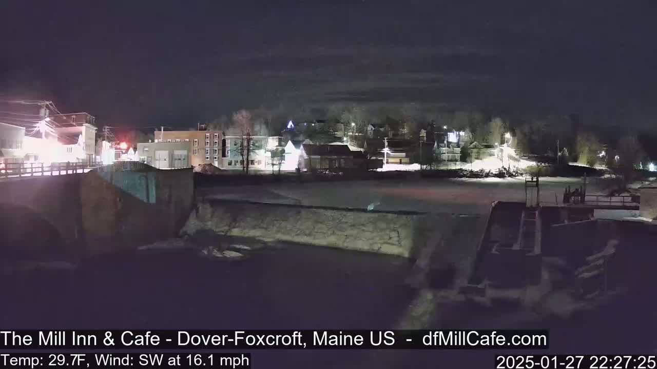 A nighttime view of a town with a river, a dam, and snow-covered ground under a partly cloudy sky.