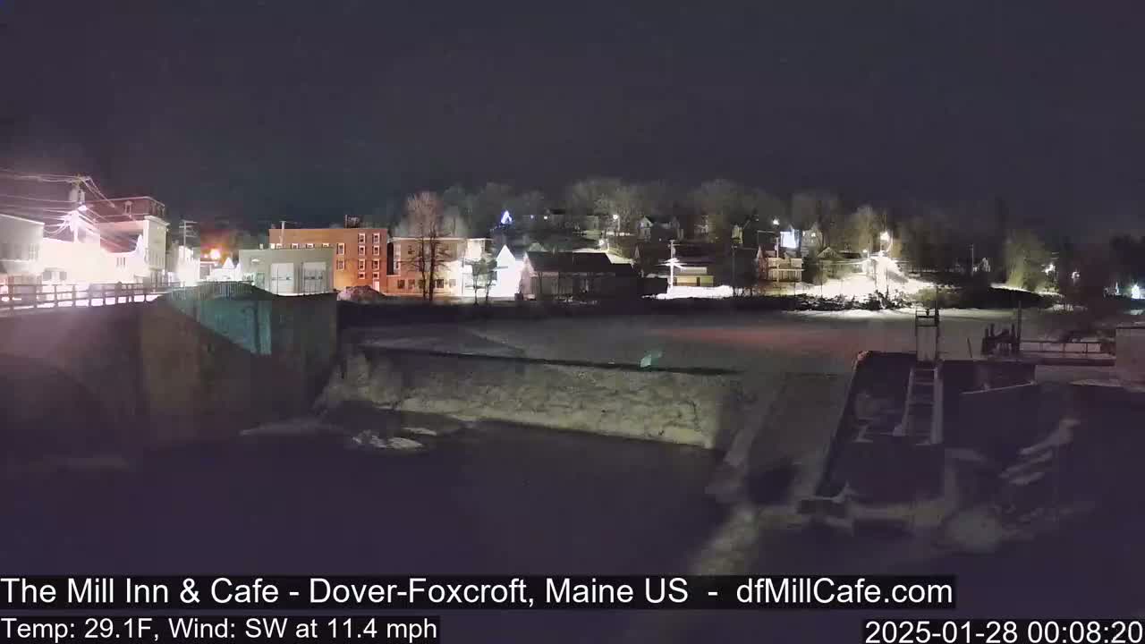 A nighttime view of a snow-covered town beside a river, with a dam and a bridge visible, under a dark sky with a southwest wind.