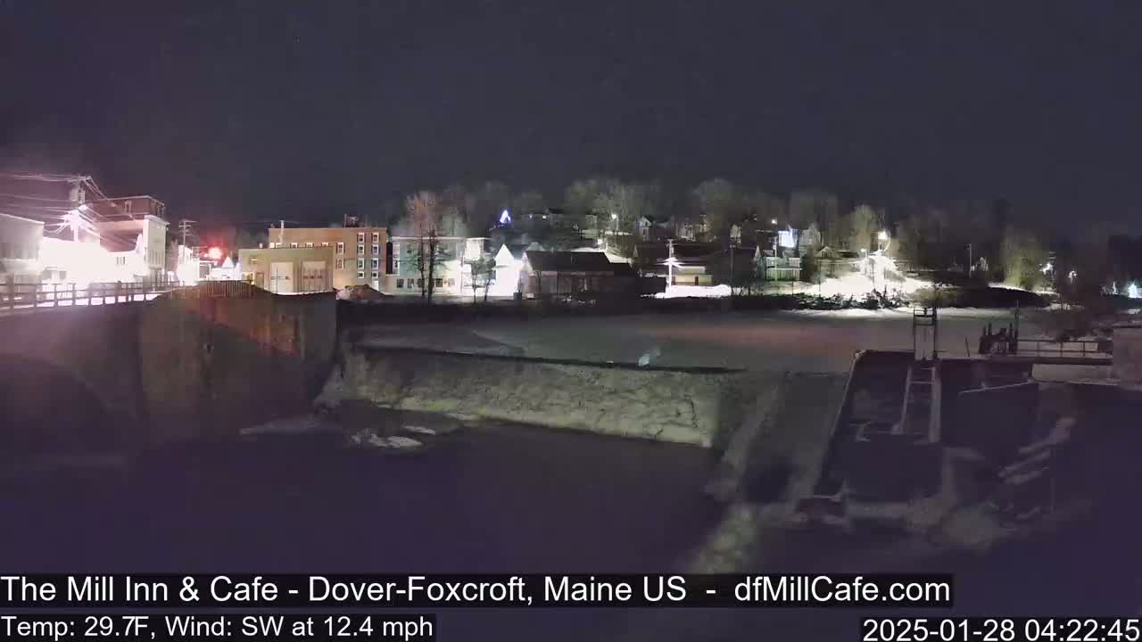 A nighttime view of a snow-covered town beside a river with a dam, under a clear, dark sky.