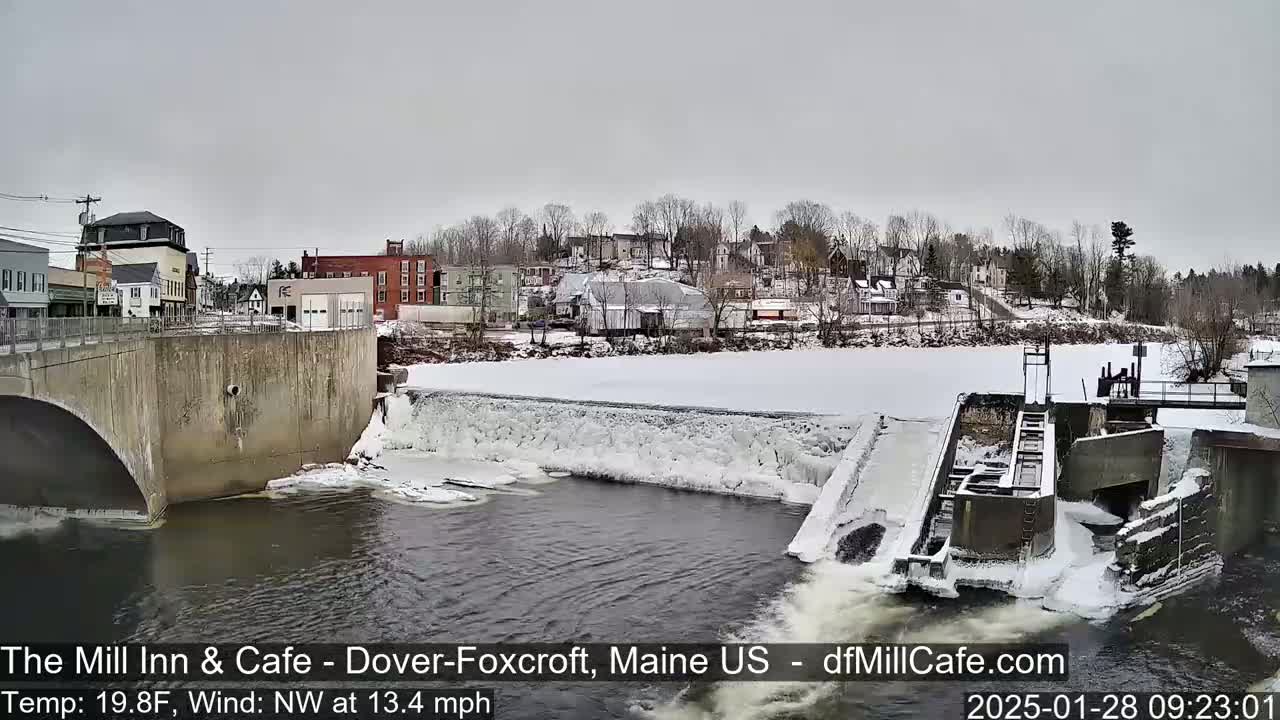 A partially frozen river flows past a dam and under a bridge, with a snow-covered town visible in the background under an overcast sky.