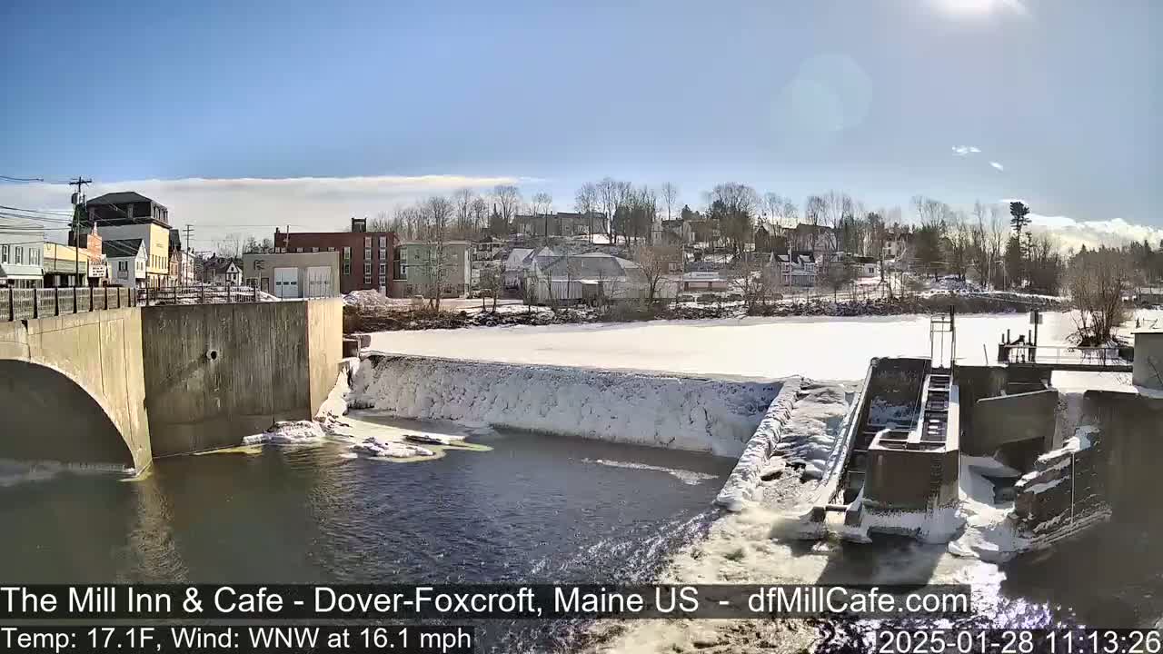 A partially frozen river flows past a dam and a small town under a sunny sky.