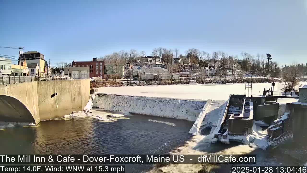 A river flows past a dam and under a bridge in a town on a sunny, cold day with snow and ice on the ground and dam.
