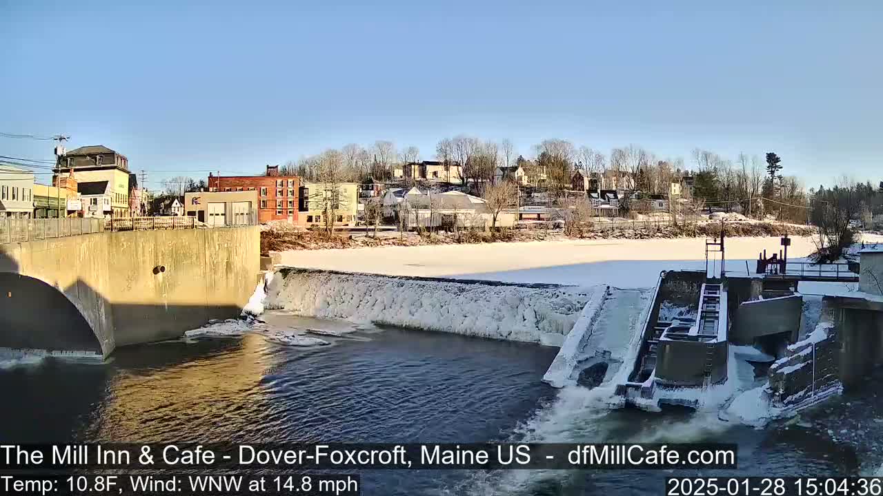 A partially frozen river flows past a dam and a small town under a clear, sunny sky.