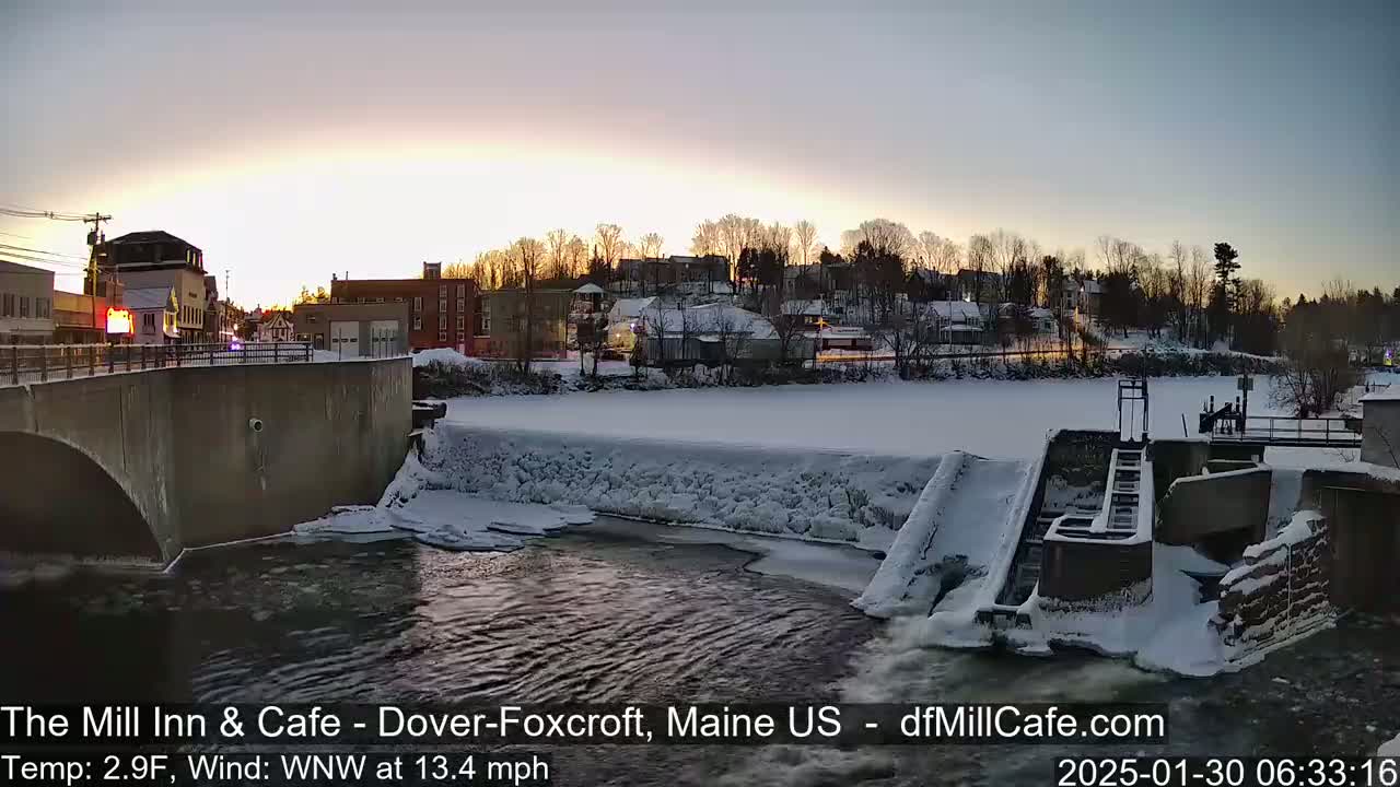 A snow-covered town at sunrise is viewed from a bridge over a partially frozen river with a dam visible in the foreground.