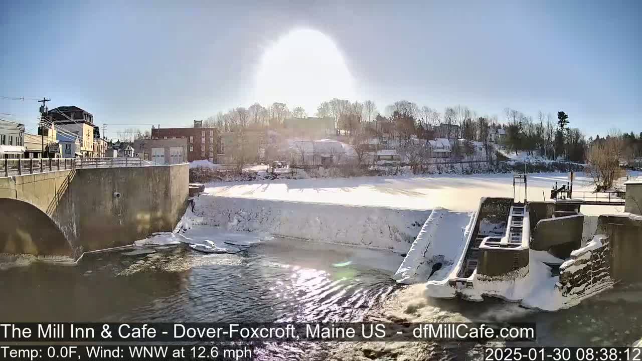 A snowy, sunny scene shows a river flowing past a dam and a small town in the background under a bright, clear sky.