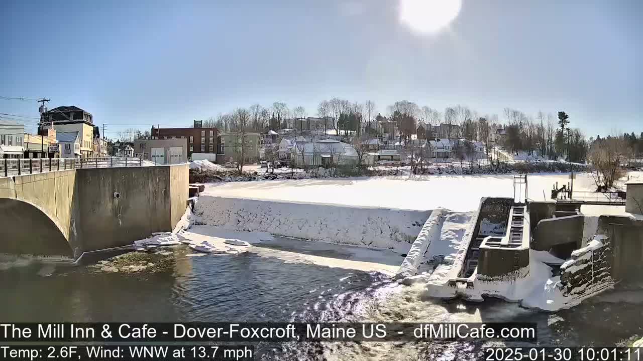 A sunny, cold day shows a snow-covered dam and river, with a small town visible in the background.